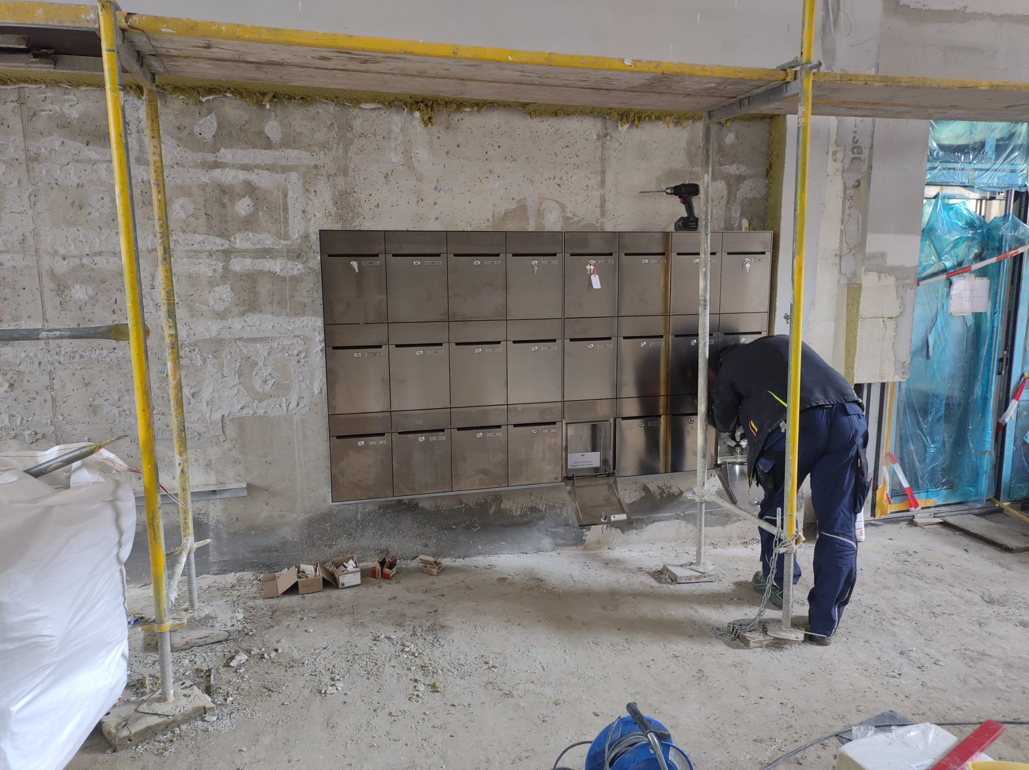 Montaż skrzynek pocztowych ze stali nierdzewnej na surowej ścianie wewnątrz budynku, widoczne rusztowanie i materiały budowlane, pracownik pochylony nad instalacją.
