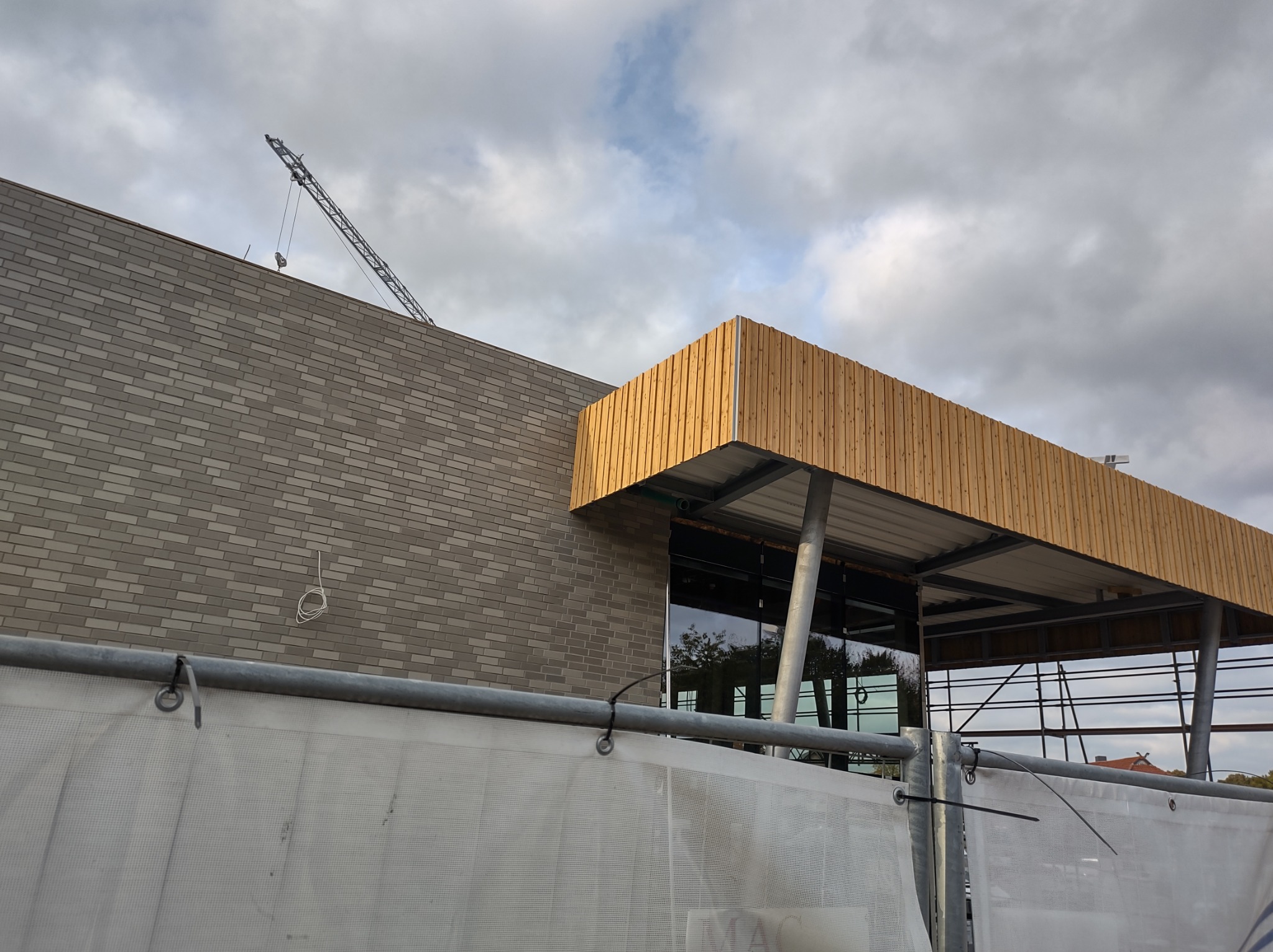 Wznoszony budynek z elewacją z szarej cegły i drewnianym zadaszeniem, widoczny żuraw budowlany na dachu, zabezpieczenie budowy w postaci białej siatki na pierwszym planie.