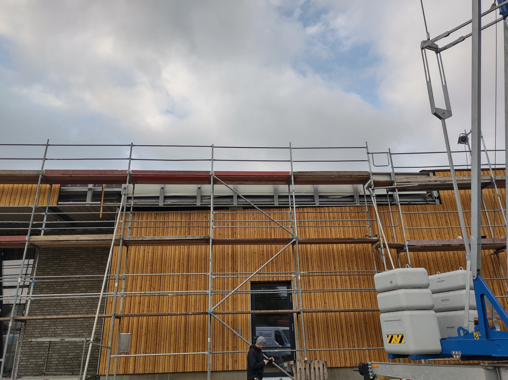 Montaż drewnianej elewacji z wykorzystaniem rusztowania i podnośnika, widoczny pracownik w trakcie wykonywania prac na wysokości.
