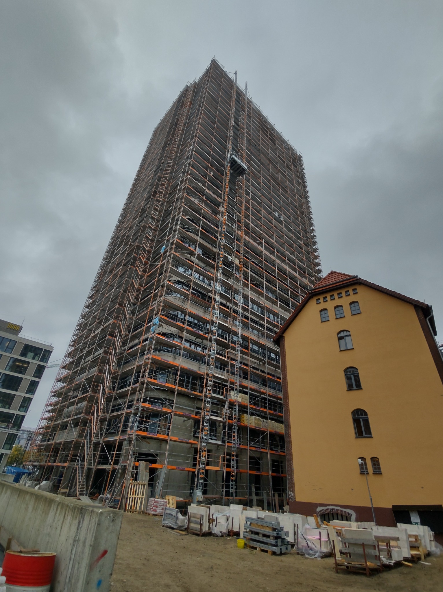 Wysoki budynek w trakcie prac elewacyjnych, całkowicie otoczony rusztowaniami, z widoczną windą budowlaną, na tle pochmurnego nieba i sąsiedniego budynku o żółtej elewacji.