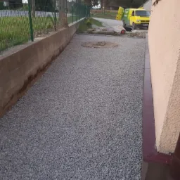 Wysypany szary żwir na podwórku przy budynku, widoczny fragment fundamentu i ogrodzenie, w tle żółty samochód dostawczy z otwartymi drzwiami.