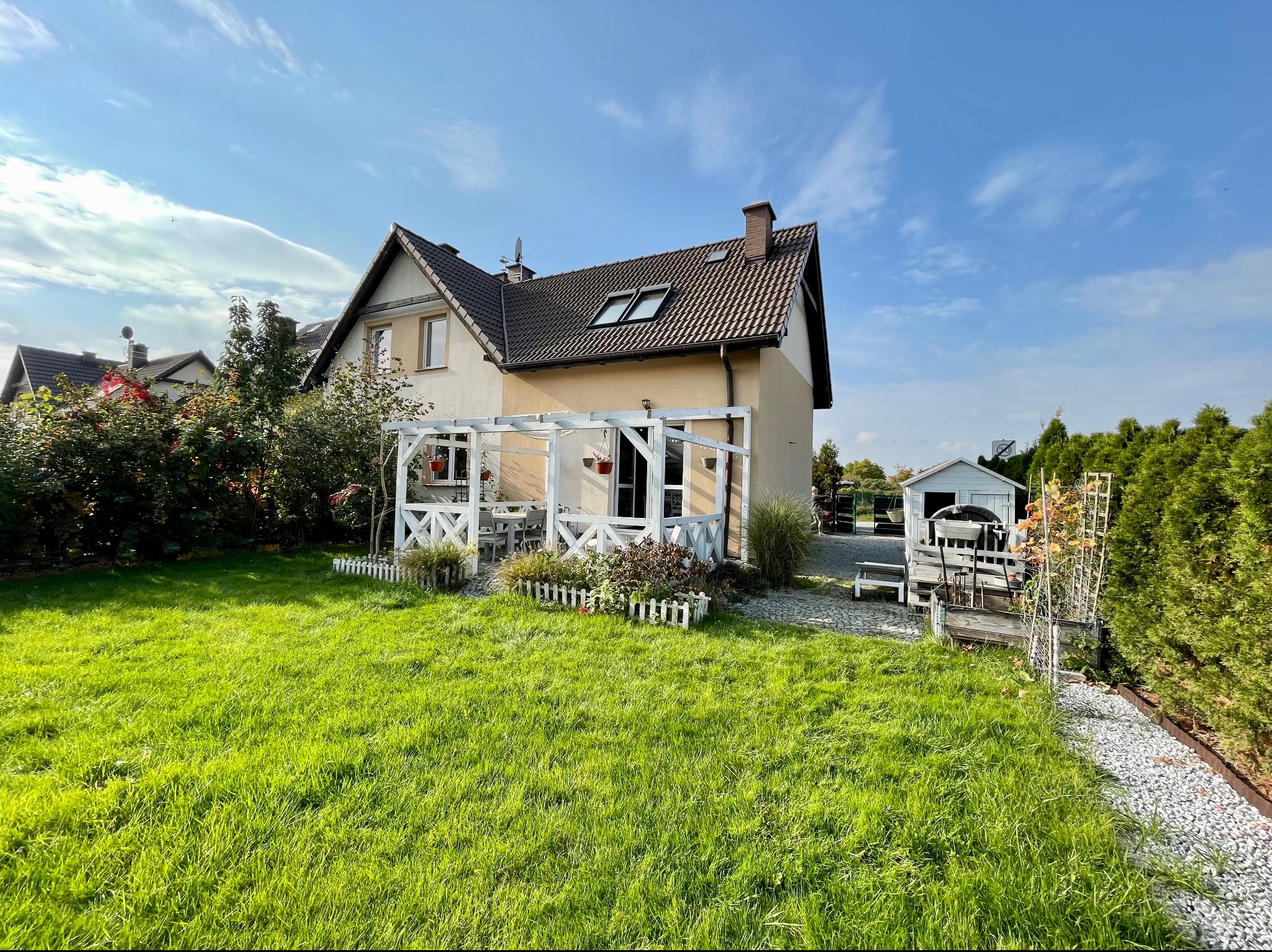 Dom jednorodzinny z ogrodem, widoczny z perspektywy trawnika, z białym, drewnianym tarasem i pergolą, z elementami małej architektury ogrodowej. W tle błękitne niebo.