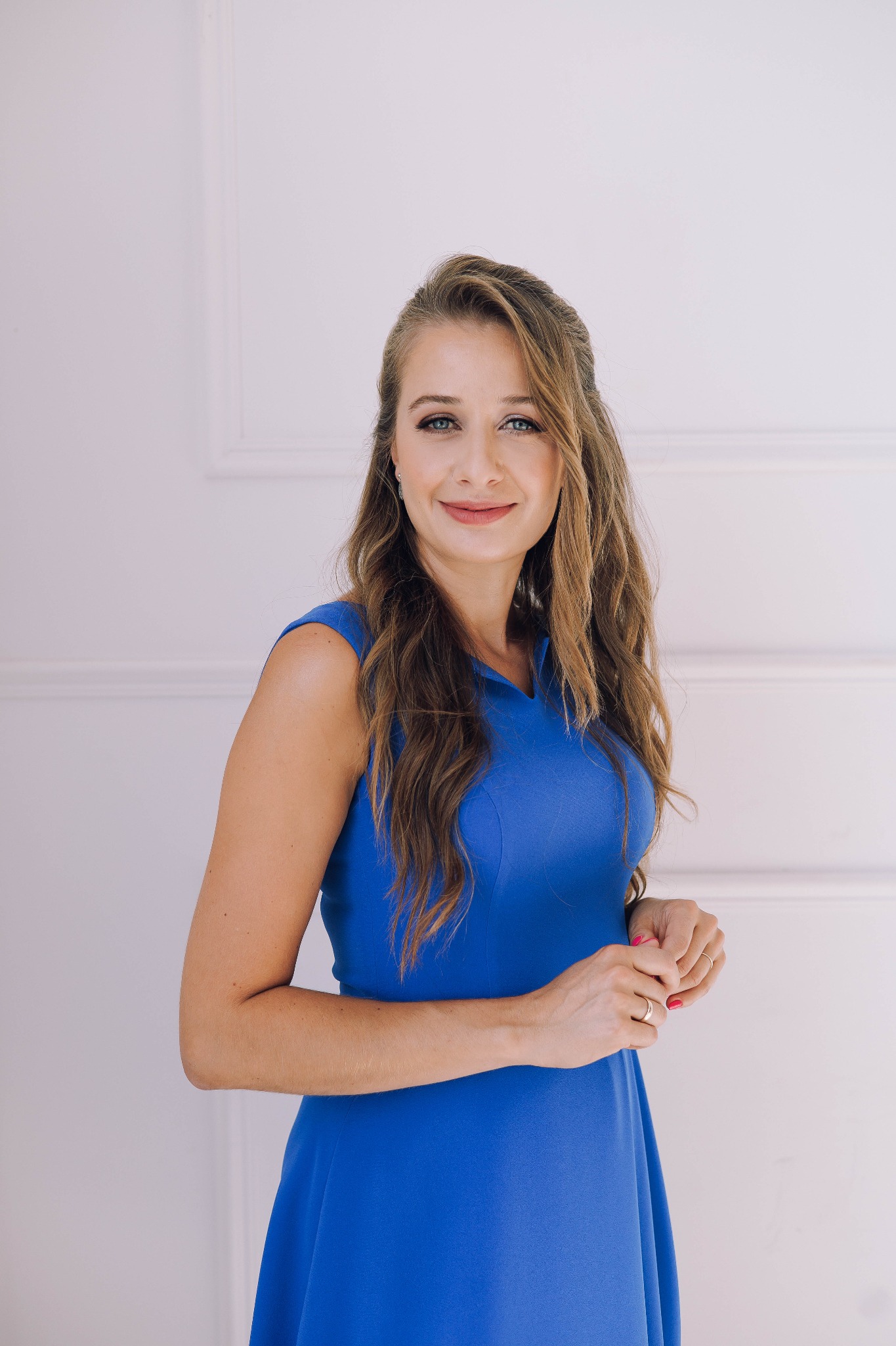 Portret uśmiechniętej kobiety w niebieskiej sukience na tle białej ściany z delikatnymi zdobieniami.