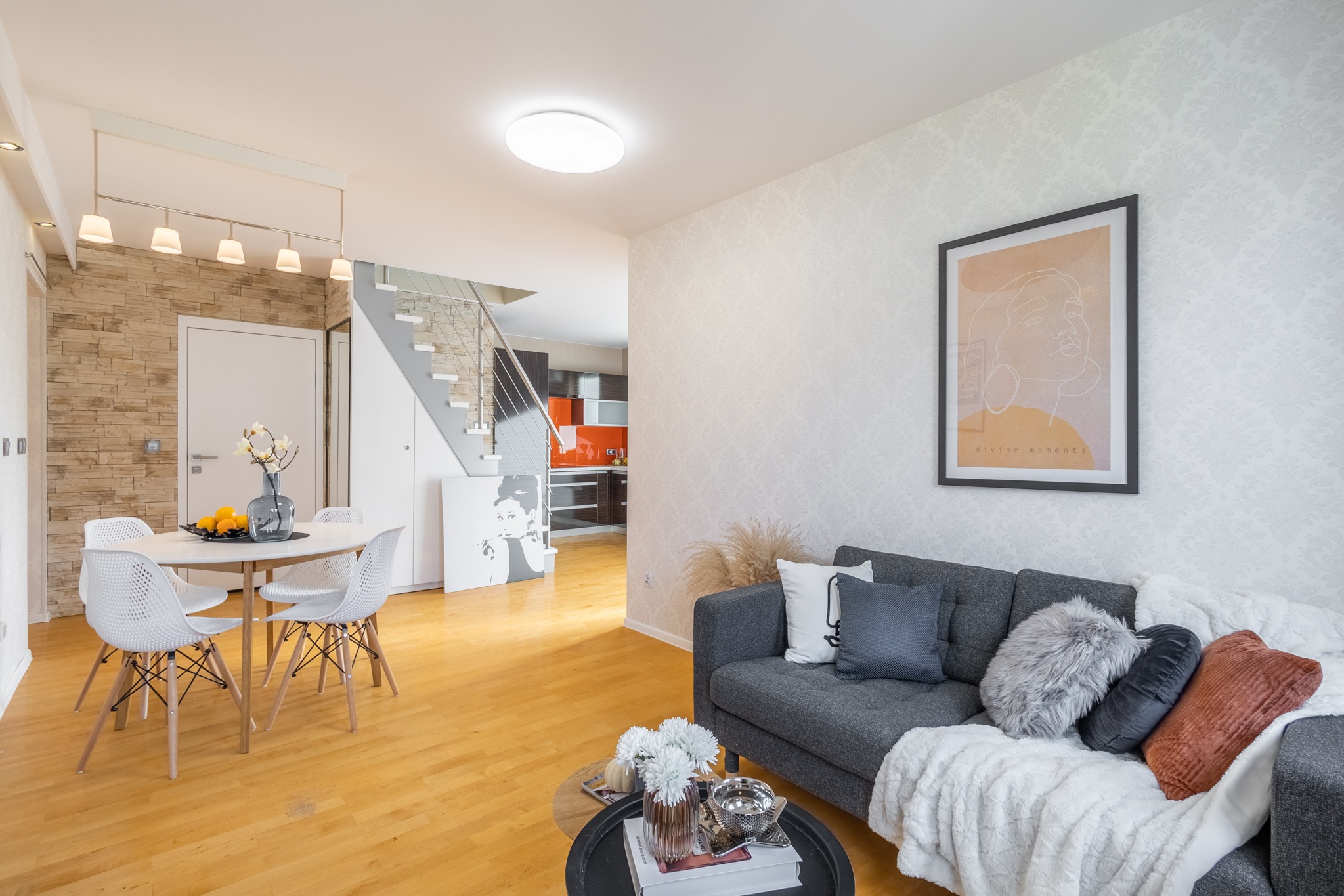 Jasne wnętrze domu z drewnianą podłogą, szarym narożnikiem z poduszkami, okrągłym stołem z krzesłami i schodami prowadzącymi na piętro, widok na kuchnię w tle.