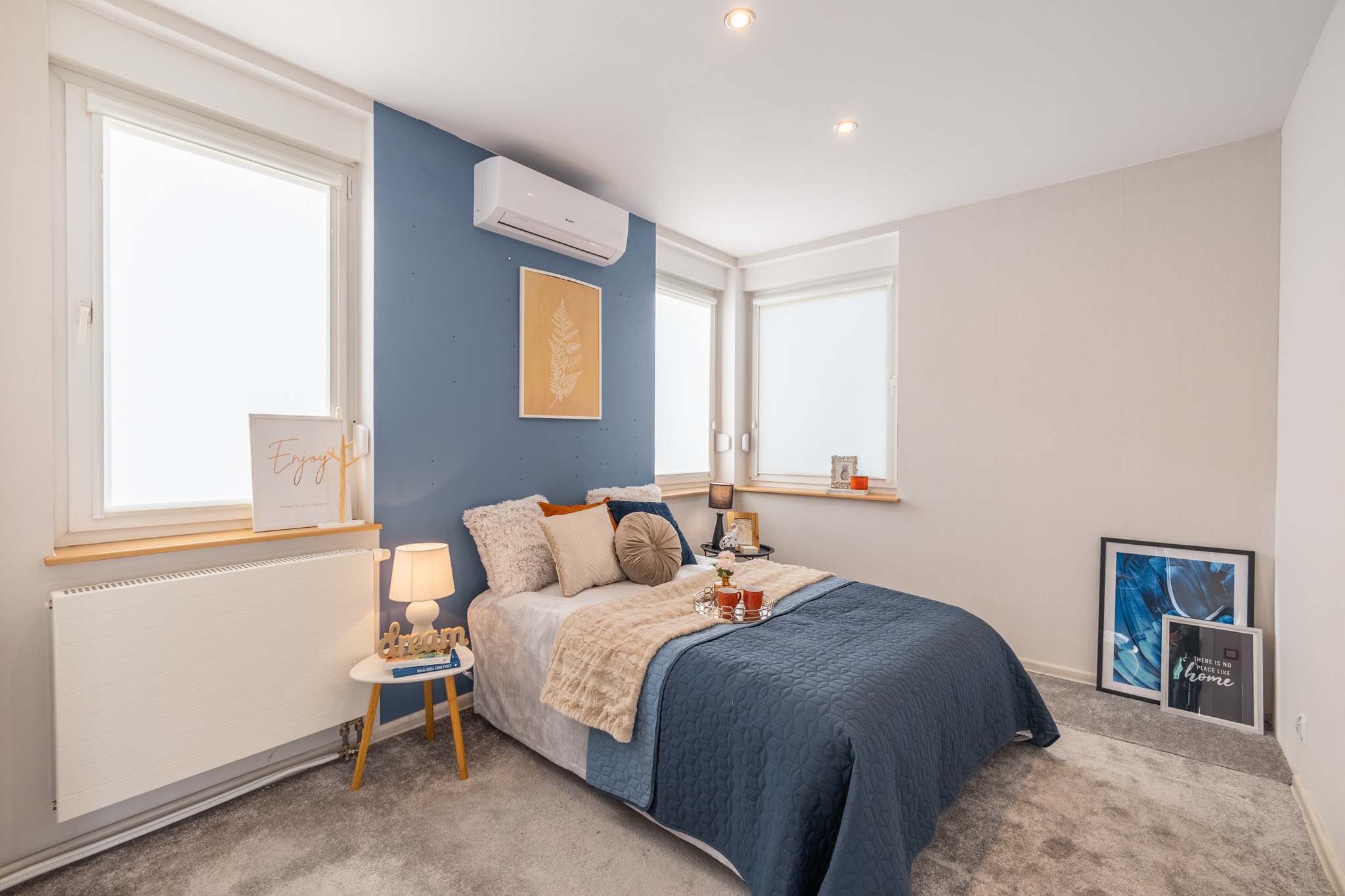 Jasna sypialnia z niebieską ścianą, klimatyzacją, łóżkiem z dekoracyjnymi poduszkami, stolikiem nocnym z lampką i dekoracjami okiennymi.