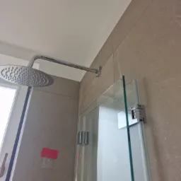 Nowoczesna kabina prysznicowa z deszczownicą, jasne płytki na ścianach, widoczne zawiasy i uszczelki kabiny, fragment okna po lewej stronie i czerwona kartka przyklejona do ściany.