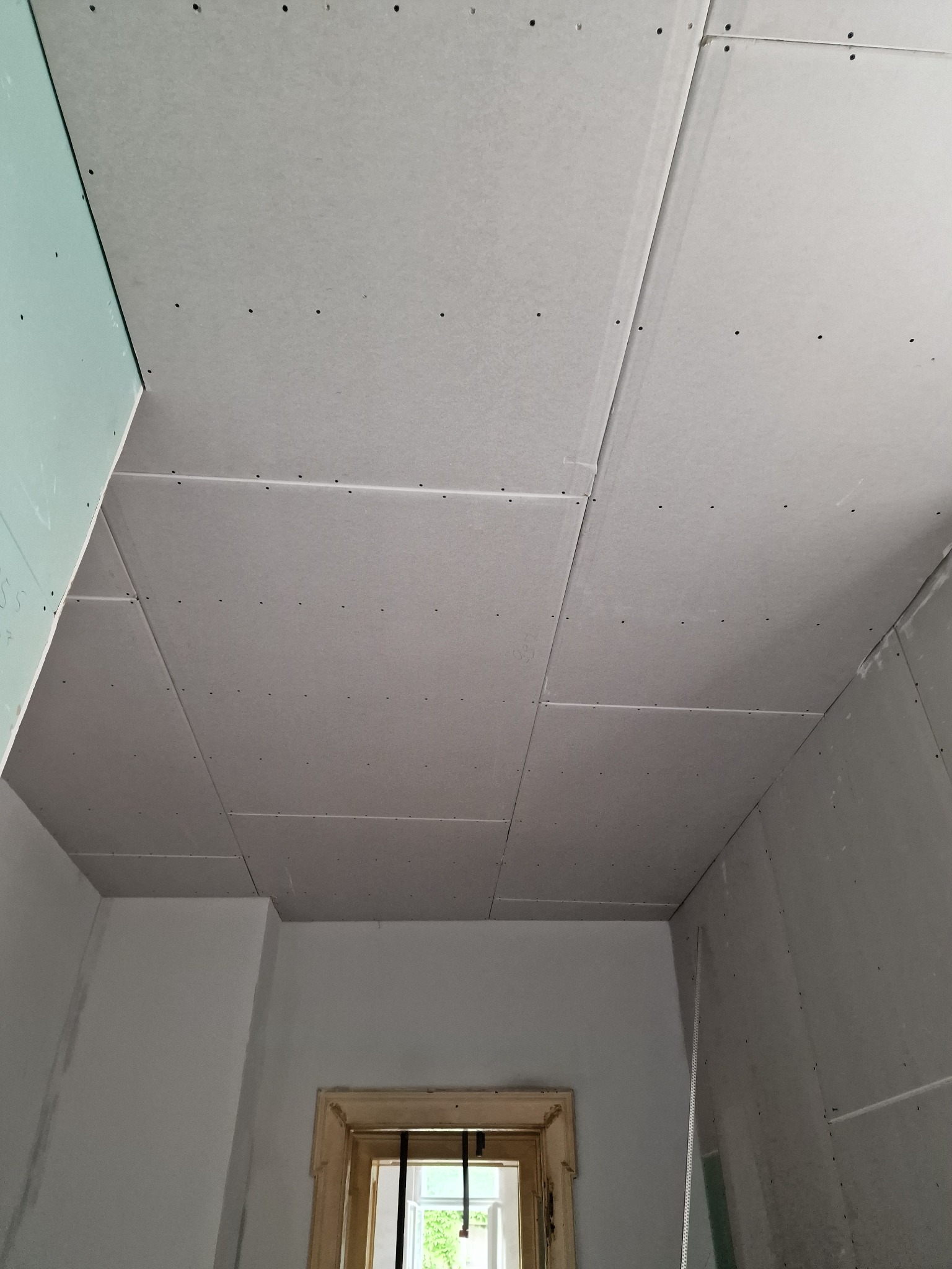 Montaż płyt gipsowo-kartonowych na suficie pod skosem, widoczne wkręty i łączenia płyt, fragment ściany z oknem w tle.