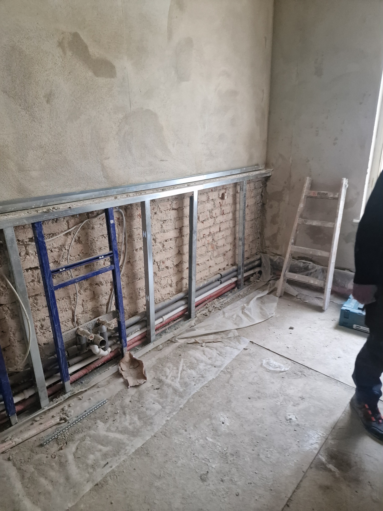 Ściana w trakcie remontu z widoczną konstrukcją pod zabudowę, instalacją hydrauliczną, starą cegłą i fragmentem drabiny.