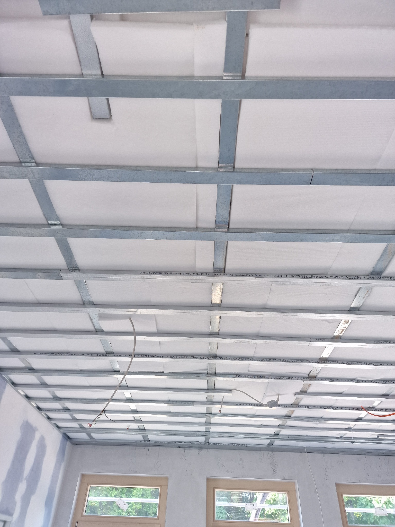 Montaż metalowej konstrukcji pod sufitem podwieszanym z widoczną izolacją w pomieszczeniu w Skawie, z oknami w tle.