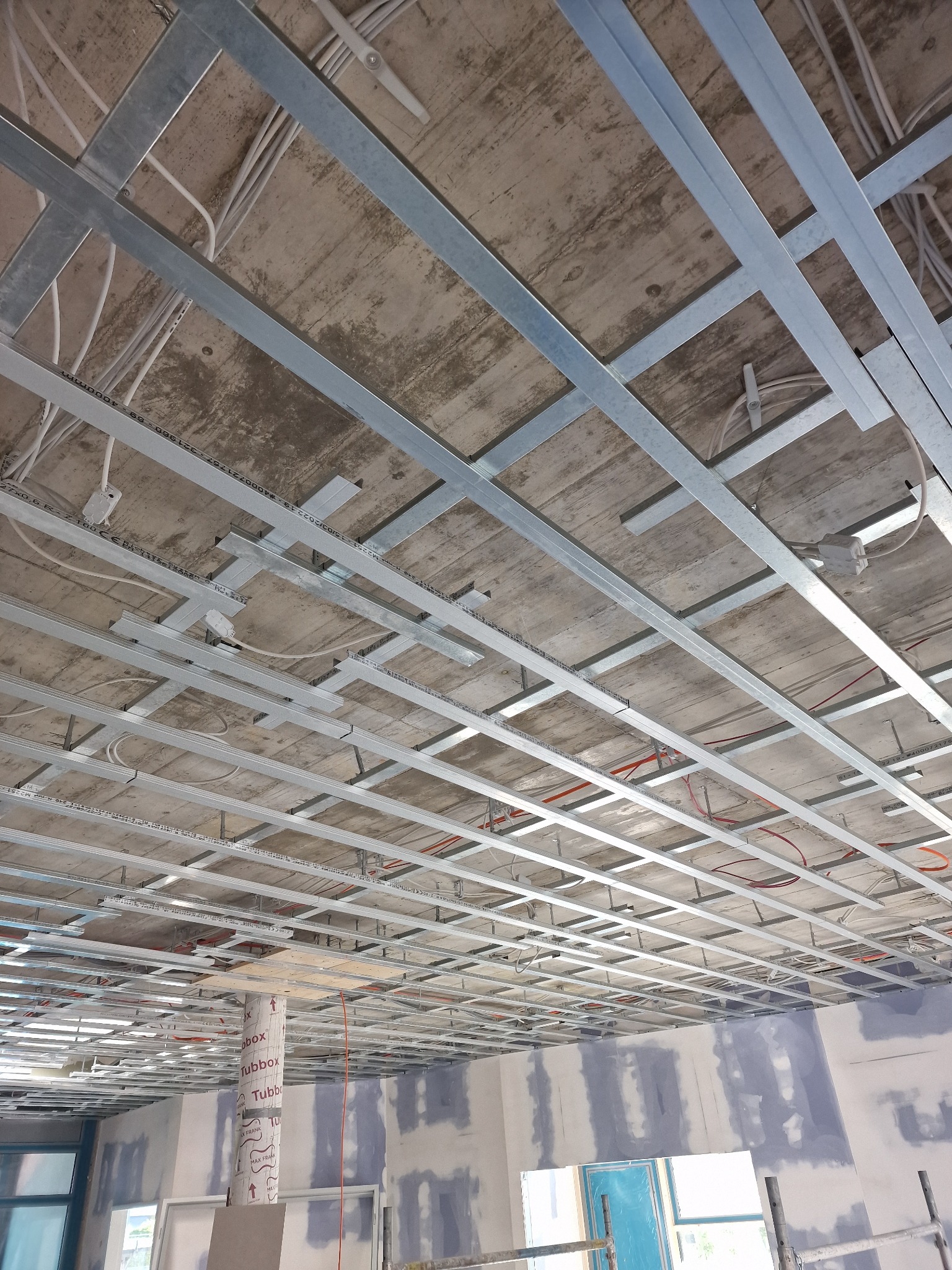 Konstrukcja metalowa podwieszanego sufitu na tle surowego betonowego stropu z widocznymi przewodami elektrycznymi, ściany w trakcie szpachlowania, tuby ochronne na słupach.