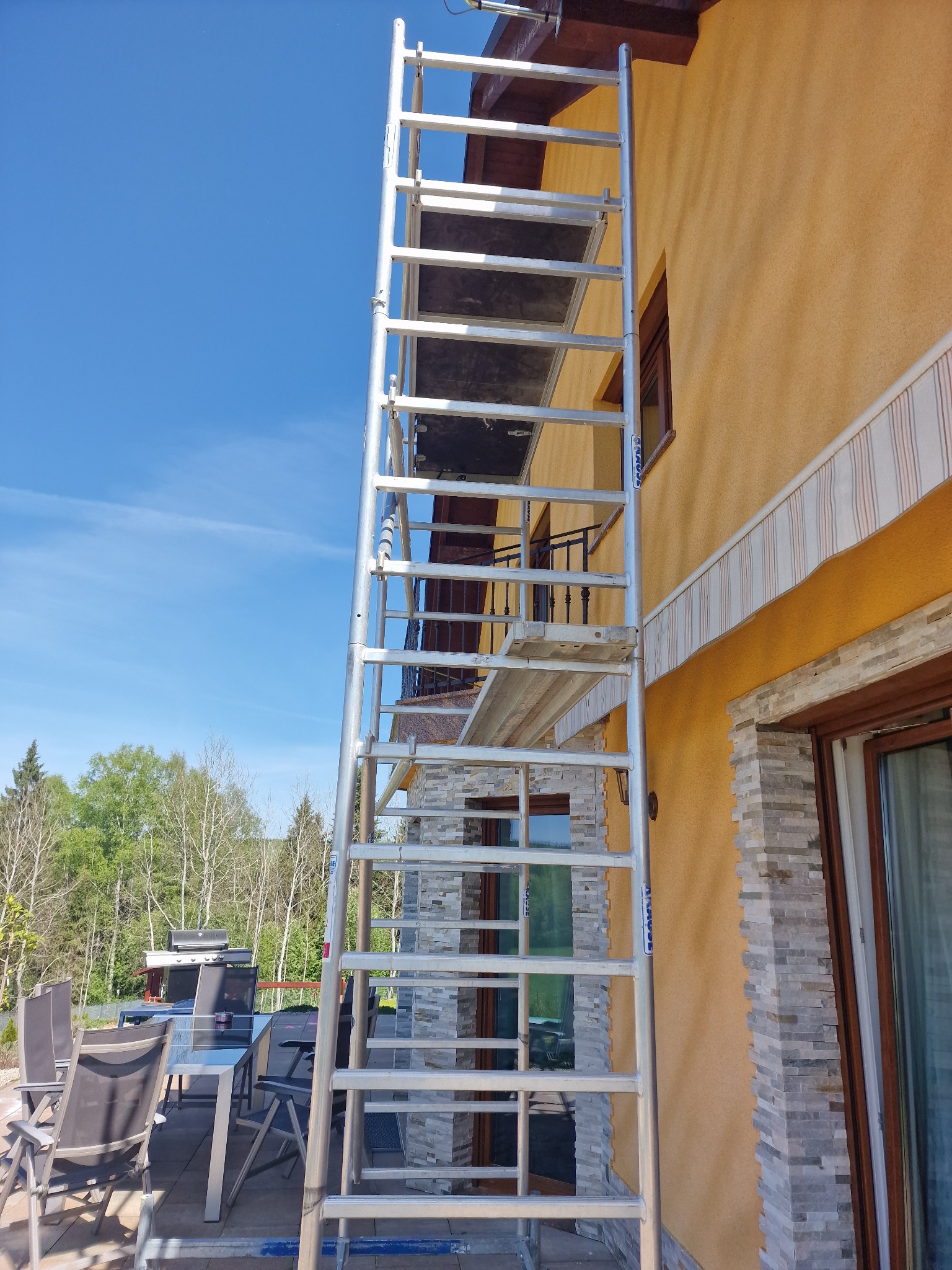 Wysoka aluminiowa wieża rusztowania stoi przy pomarańczowej ścianie domu z dekoracyjnymi kamiennymi elementami. W tle widoczny taras z meblami ogrodowymi i drzewa.