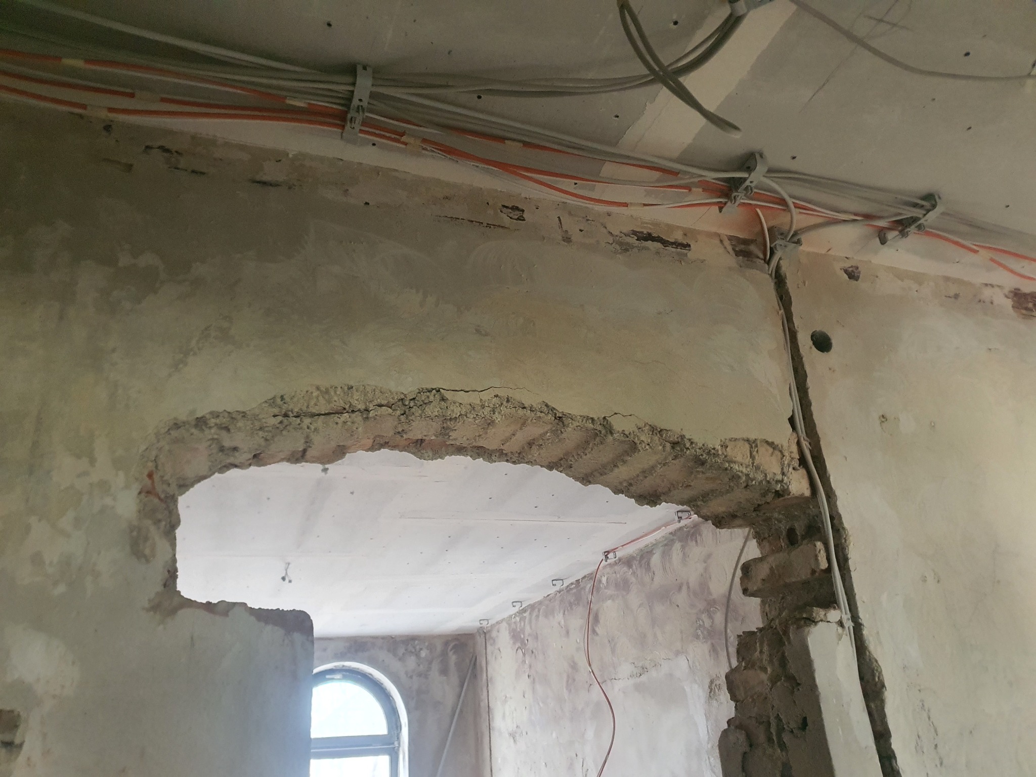 Demontaż fragmentu ściany z odsłoniętą cegłą i instalacją elektryczną w trakcie remontu. Widoczne przewody przymocowane do sufitu oraz fragment okna w tle.