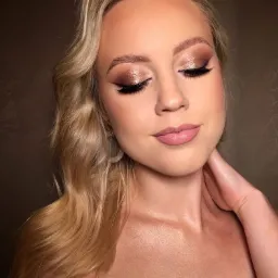 Portret kobiety z zamkniętymi oczami, prezentującej wieczorowy, błyszczący makijaż oczu w odcieniach brązu i beżu z wyraźną kreską, delikatny róż na ustach, blond włosy ułożone w fale i subtelne...