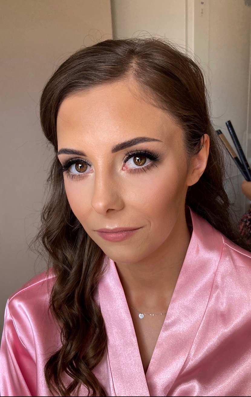Portret kobiety po makijażu ślubnym, z podkreślonymi oczami i brązowymi włosami ułożonymi w fale, ubrana w satynowy, różowy szlafrok i delikatny naszyjnik z serduszkiem.