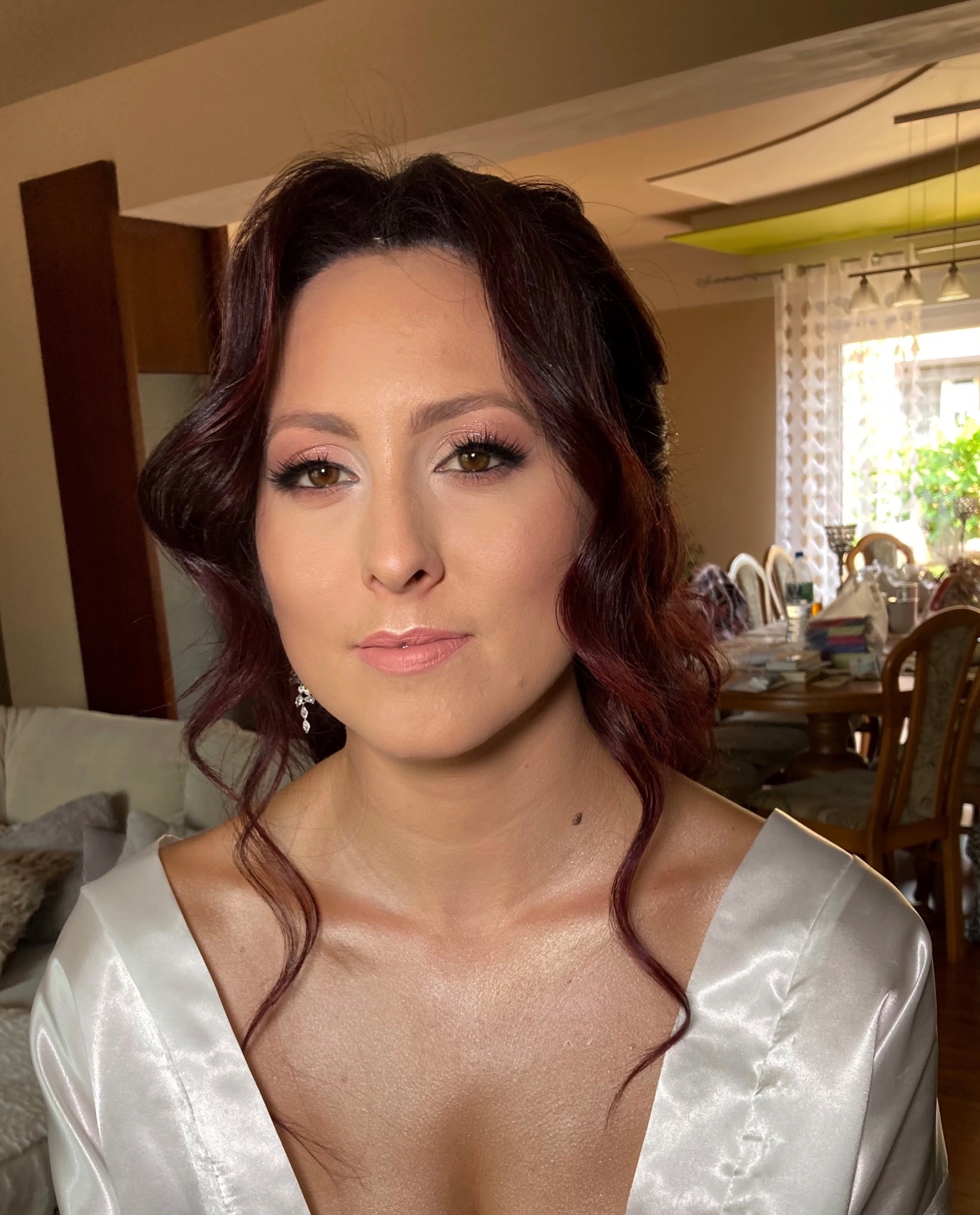 Portret kobiety z delikatnym, różowym makijażem, podkreślonymi rzęsami i upiętymi, falowanymi, ciemnymi włosami, ubrana w satynowy szlafrok, w tle widoczny salon.