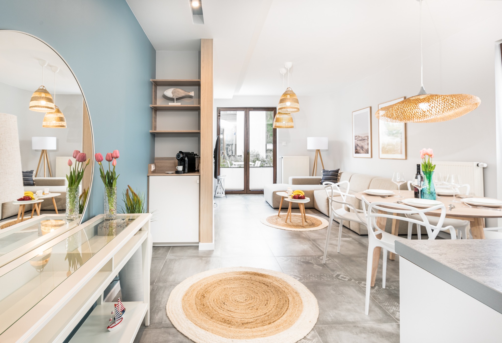 Jasne, przestronne wnętrze mieszkania z elementami wystroju w stylu skandynawskim: okrągłe dywaniki z juty, drewniane lampy, okrągłe lustro i bukiet tulipanów na stole.