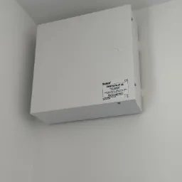 Biała skrzynka alarmowa Satel PERFECTA-IP 32 zamontowana na białej ścianie w rogu pomieszczenia, widoczne ślady po instalacji poniżej.