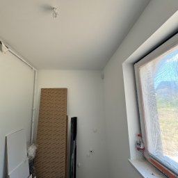 Klasa Instal Damian Klasa - Wnętrze w trakcie wykańczania, widoczne przewody wystające z sufitu, okno zabezpieczone folią, płyty meblowe oparte o ścianę, butelka wody na parapecie.