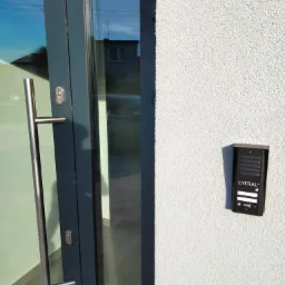 Czarny wideodomofon Cyfral zamontowany na szarej, chropowatej ścianie obok wejścia z szarymi drzwiami i długim, metalowym uchwytem.