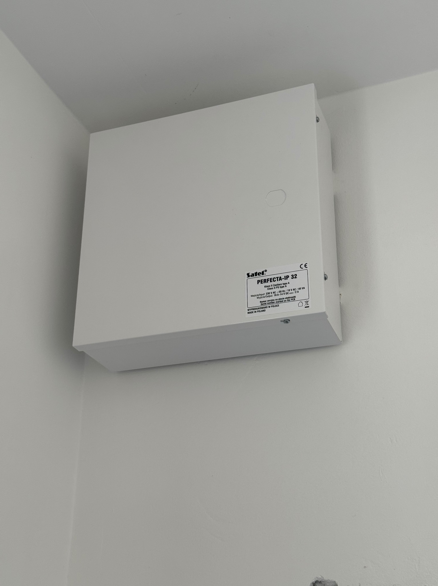 Biała skrzynka alarmowa Satel PERFECTA-IP 32 zamontowana na białej ścianie w rogu pomieszczenia, widoczne ślady po instalacji poniżej.