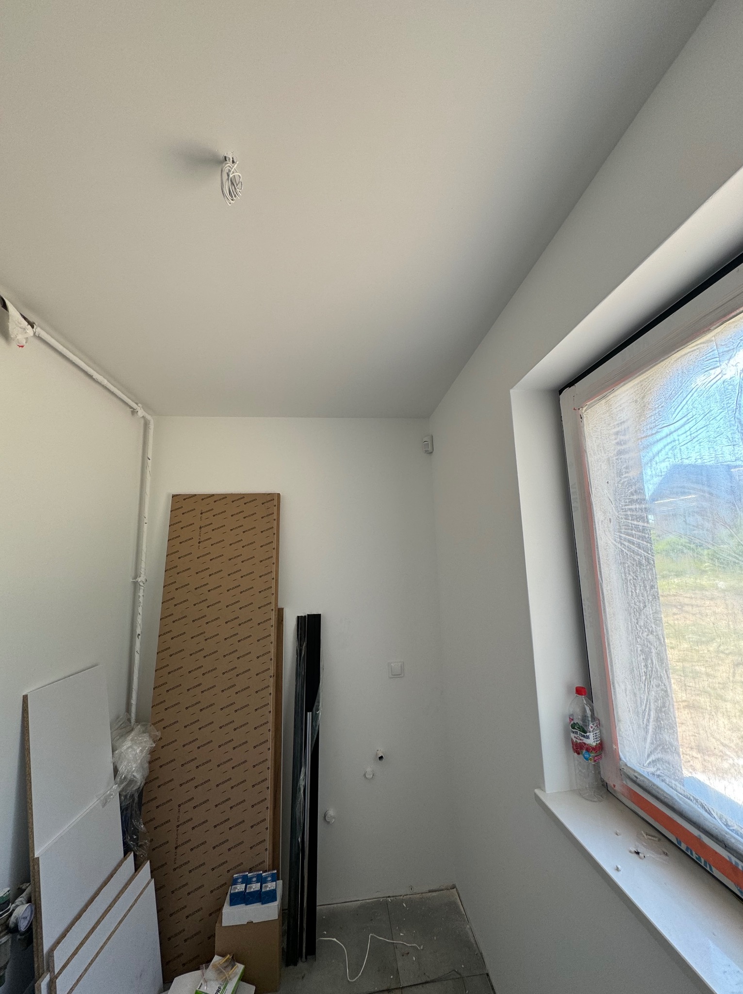 Wnętrze w trakcie wykańczania, widoczne przewody wystające z sufitu, okno zabezpieczone folią, płyty meblowe oparte o ścianę, butelka wody na parapecie.