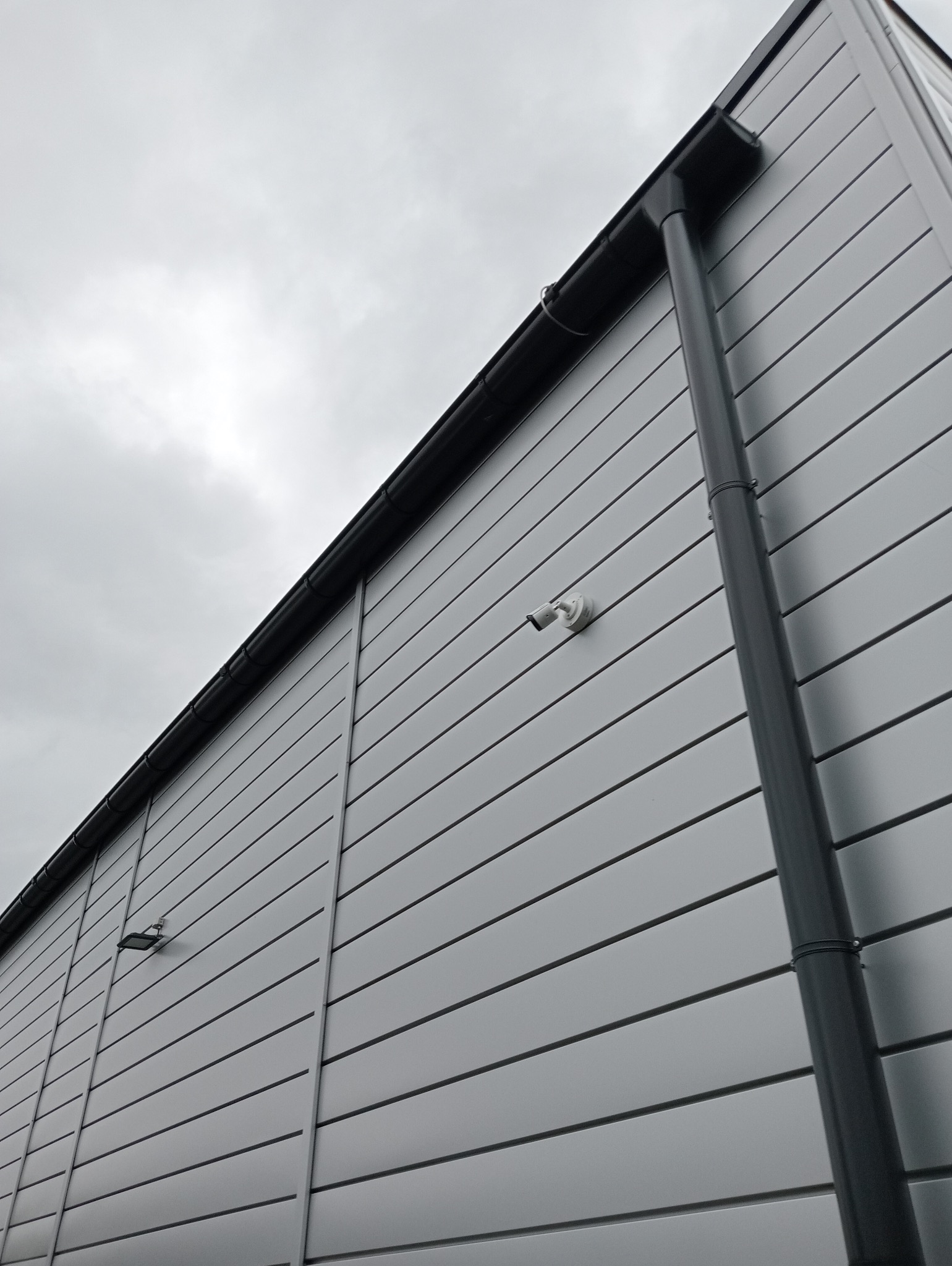 Zewnętrzna ściana budynku przemysłowego z metalowym sidingiem, na której zamontowano dwie białe kamery monitoringu. Widoczna rynna spustowa i pochmurne niebo w tle.