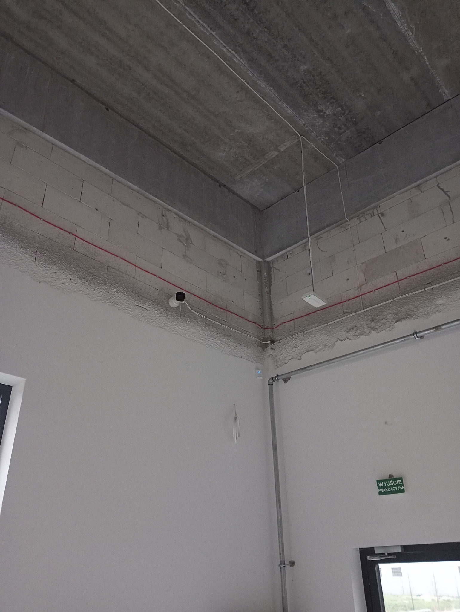 Kamera monitoringu zamontowana w rogu surowego pomieszczenia z widocznymi przewodami elektrycznymi i rurami instalacyjnymi.