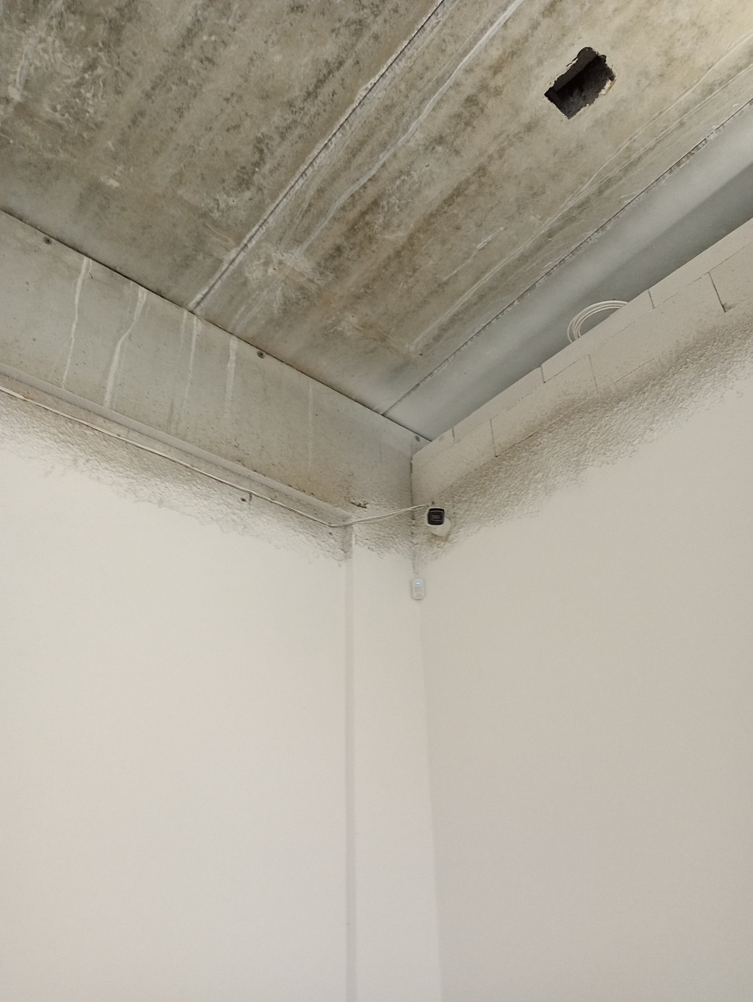 Kamera monitoringu zamontowana w rogu pomieszczenia z surowym betonowym sufitem i gładkimi, białymi ścianami.