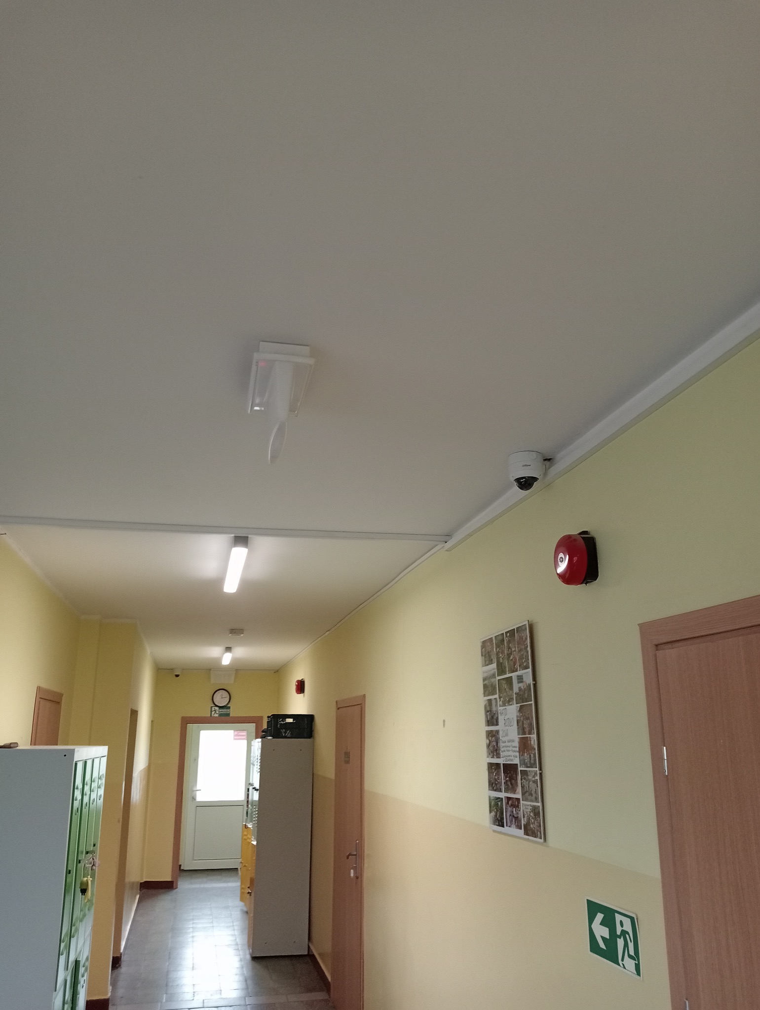 Korytarz szkolny z widoczną kamerą monitoringu, czujnikiem dymu i alarmem przeciwpożarowym na ścianie.