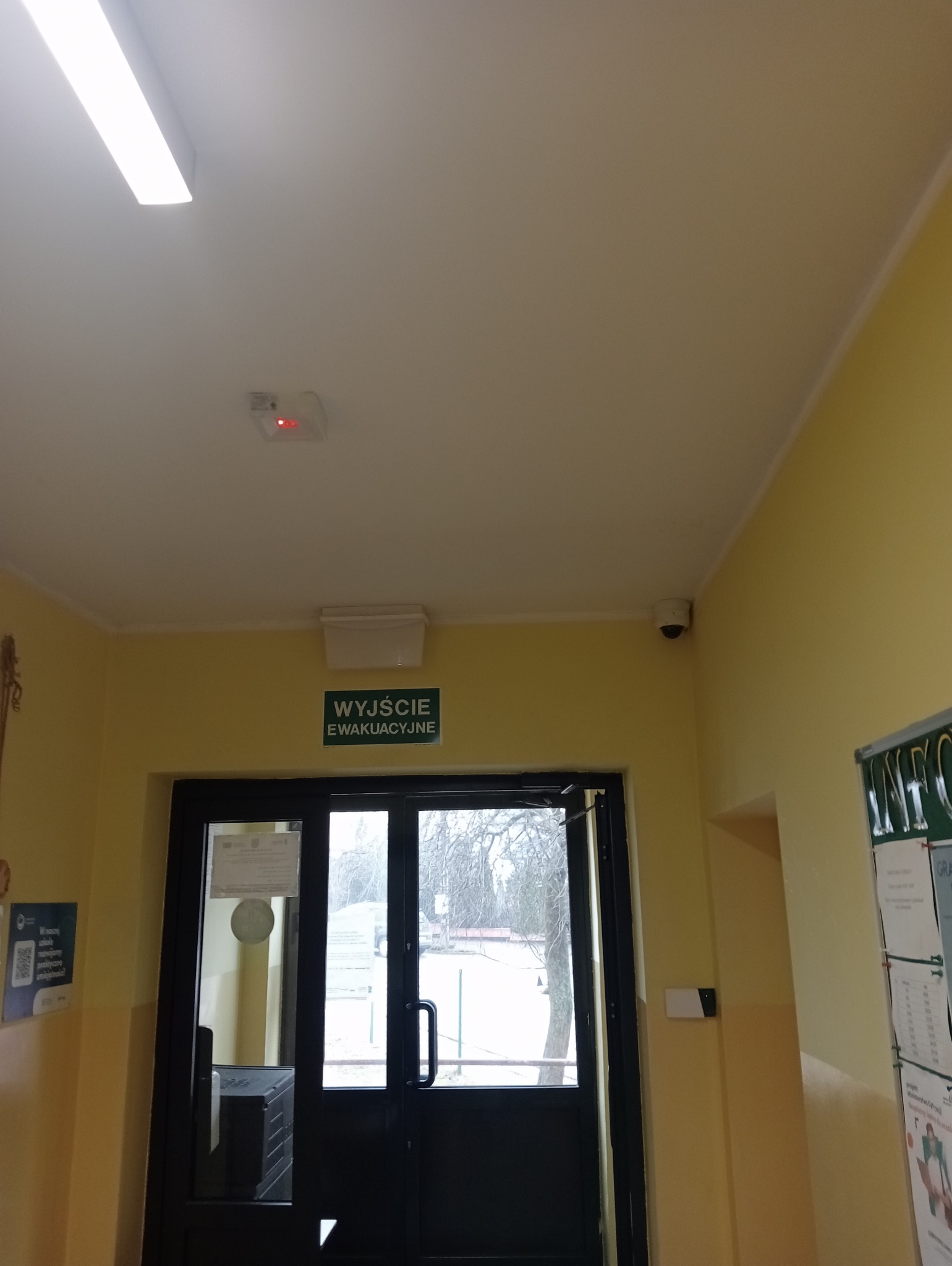 Wewnętrzny korytarz z widoczną kamerą monitoringu na suficie, czujnikiem dymu z czerwoną diodą, tabliczką informacyjną 'Wyjście Ewakuacyjne' nad czarnymi drzwiami oraz tablicą ogłoszeń po prawej...