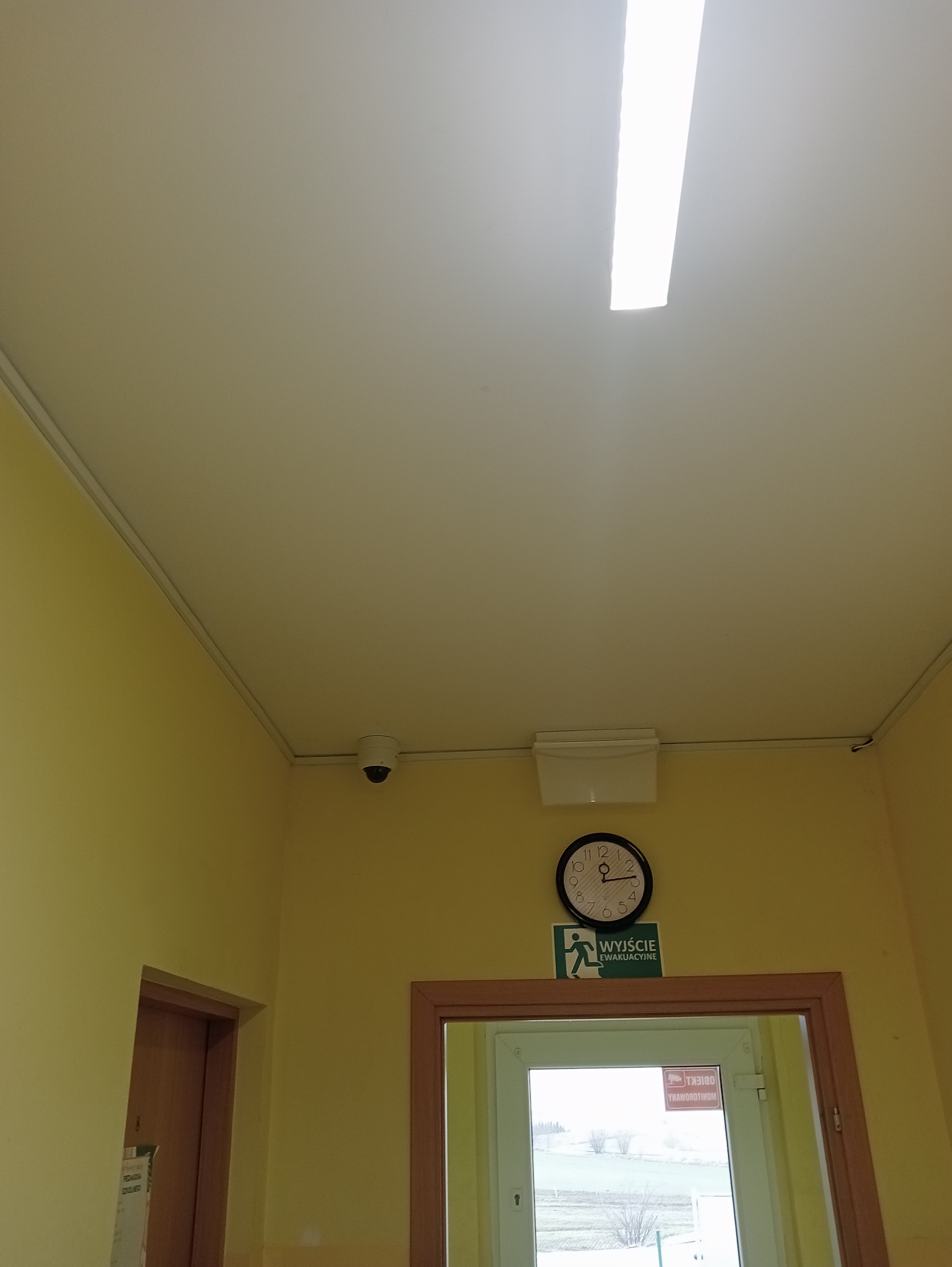 Kamera monitoringu kopułkowa zamontowana na suficie w korytarzu biurowym, widoczny zegar i tabliczka 'Wyjście Ewakuacyjne'.