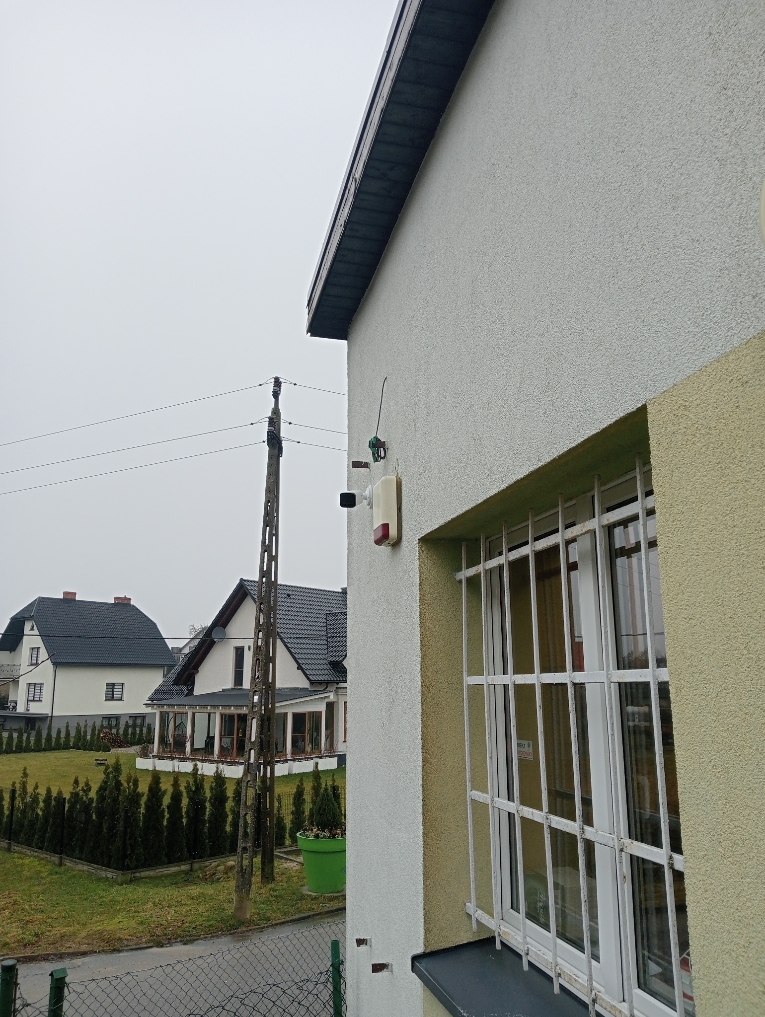Zewnętrzny czujnik alarmowy z kamerą i sygnalizatorem dźwiękowym zamontowany na elewacji domu obok okna z kratami, na tle słupa elektrycznego i innych domów.