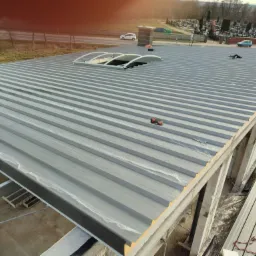 Budowa hali: Widok z góry na stalową konstrukcję dachu z świetlikiem łukowym w trakcie montażu, z pracownikiem w odblaskowej kamizelce i narzędziami na powierzchni, w tle cmentarz.