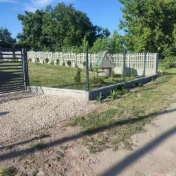 Nowoczesne ogrodzenie panelowe z bramą wjazdową, betonowa podmurówka, jasne żwirowe podłoże i studnia z drewnianą obudową w tle, widok w słoneczny dzień.