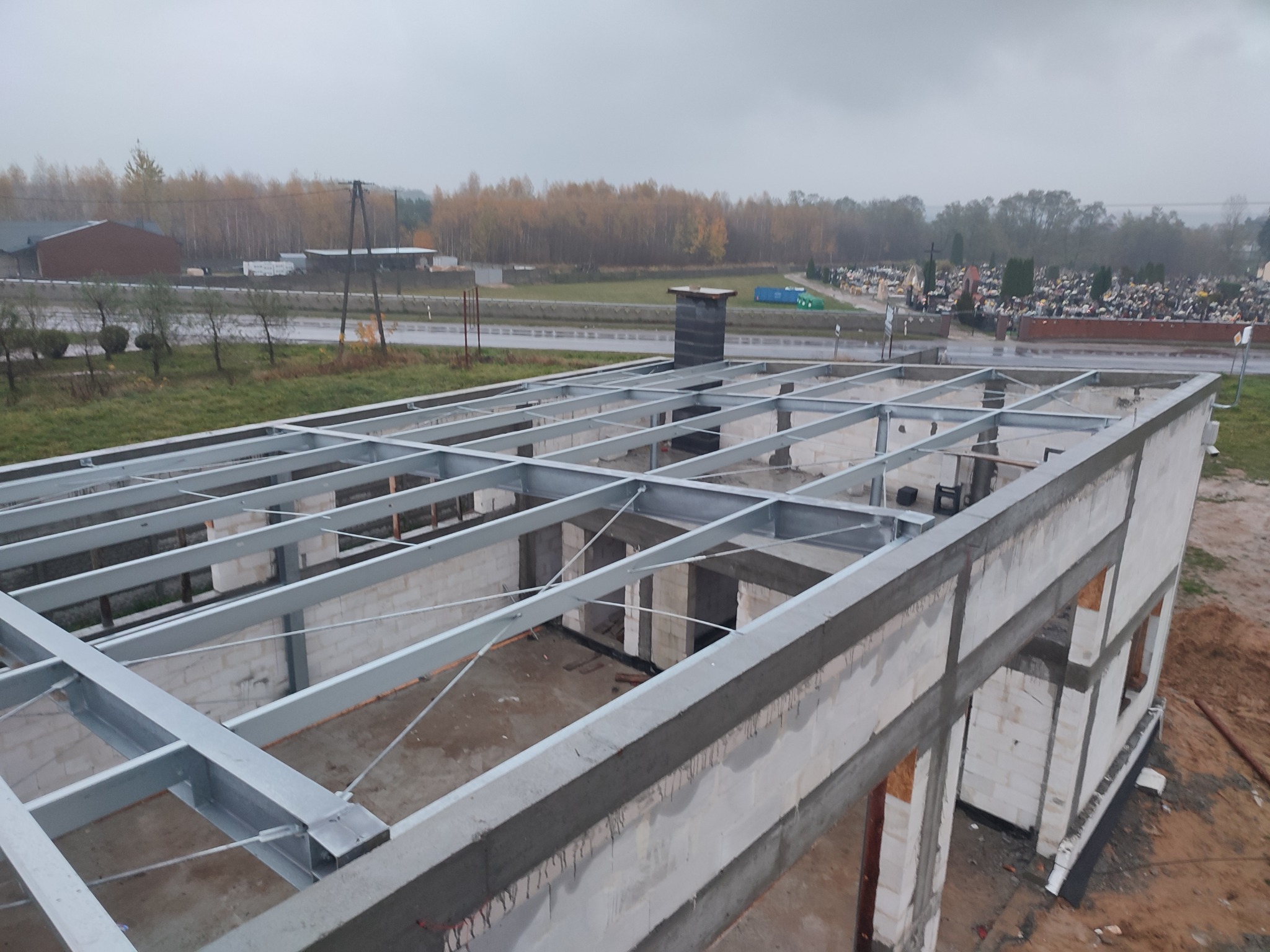 Surowy budynek w stanie surowym otwartym z widoczną konstrukcją żelbetową i stalowymi elementami dachu na tle jesiennego krajobrazu i cmentarza.