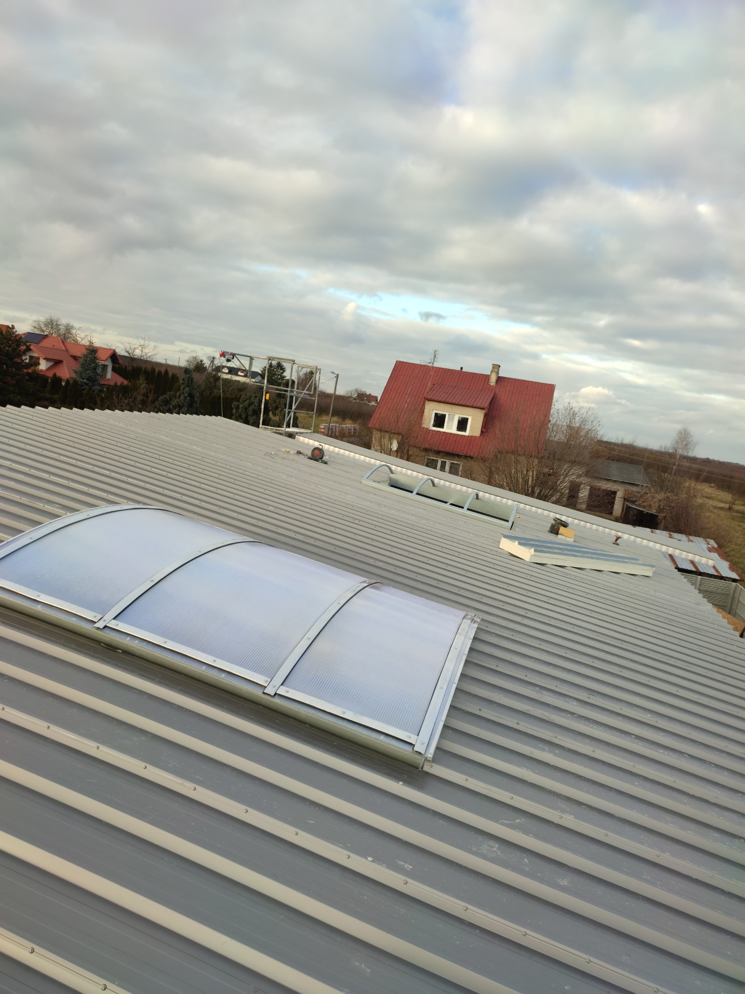 Szary dach z blachy trapezowej z zamontowanymi świetlikami dachowymi z poliwęglanu, widok z góry na tle pochmurnego nieba i zabudowań.
