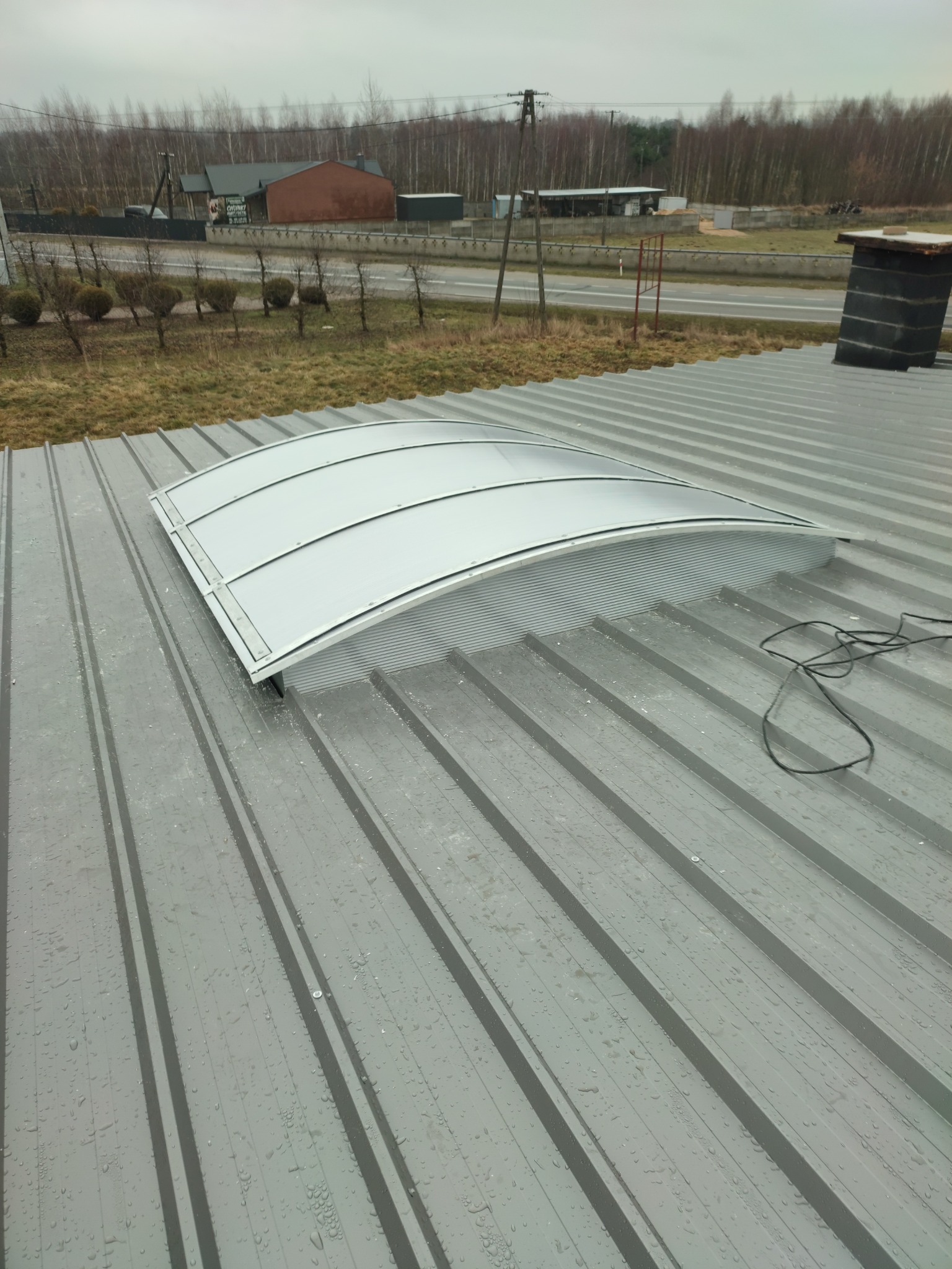 Szary dach z blachy trapezowej z zamontowanym świetlikiem łukowym, widoczne krople wody i kabel elektryczny, w tle zabudowania i drzewa.