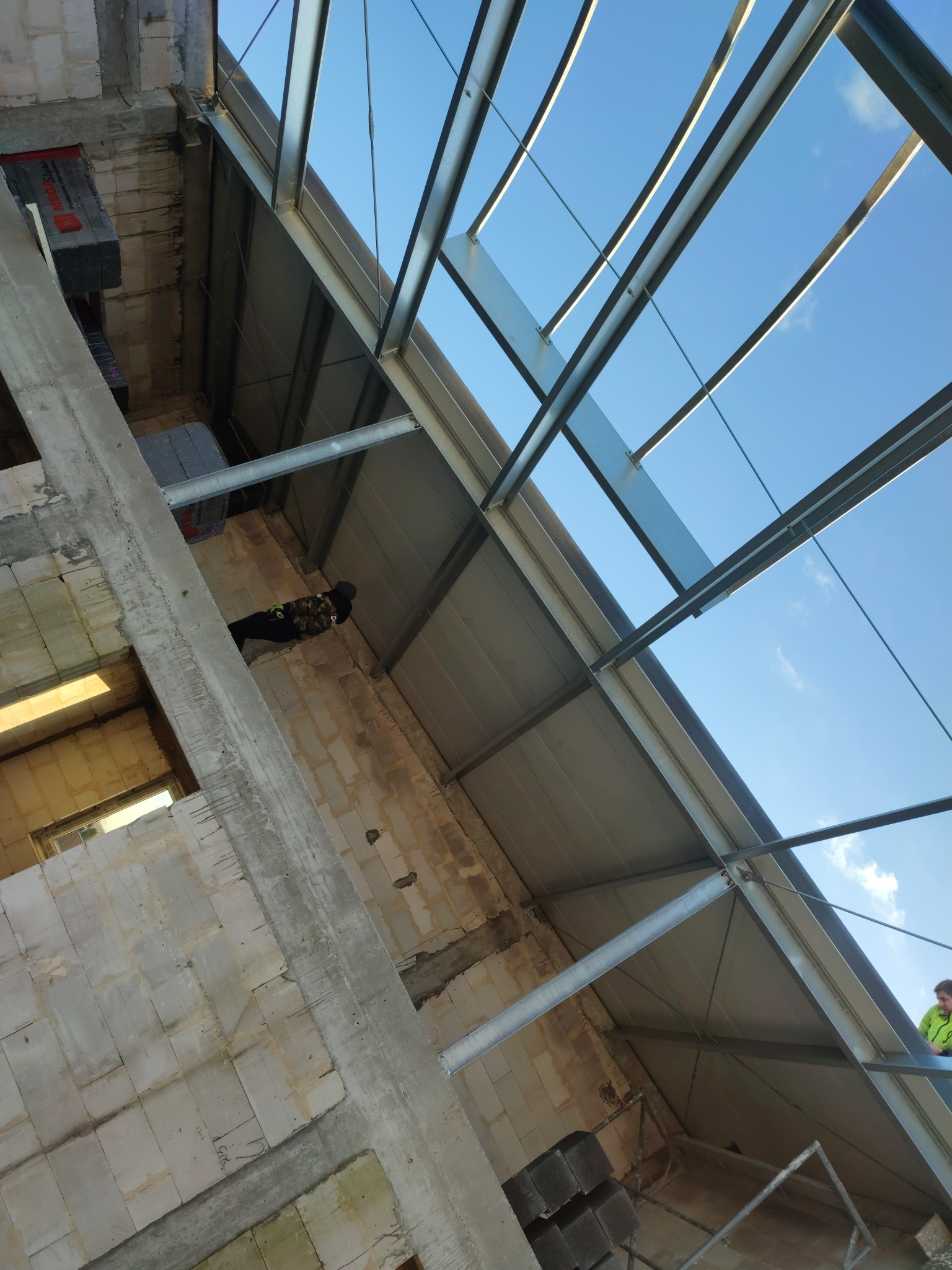 Wnętrze budowanego budynku z żelbetu i konstrukcji stalowej, widoczne ściany z bloczków, betonowy słup, stalowa konstrukcja dachu i fragment nieba.