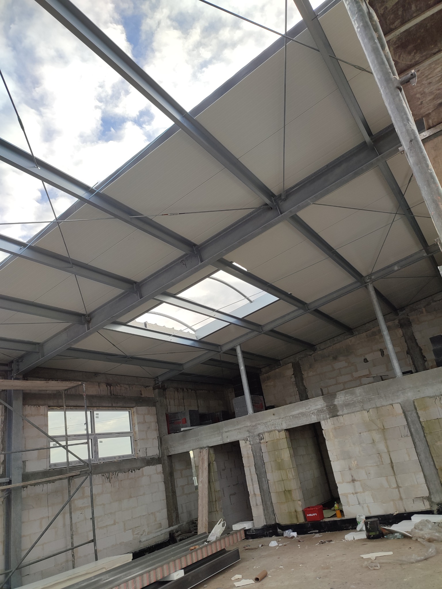 Wnętrze w trakcie budowy: stalowa konstrukcja dachu z panelami, widok na niebo przez świetlik, ściany z bloczków betonowych, rusztowania i narzędzia budowlane.