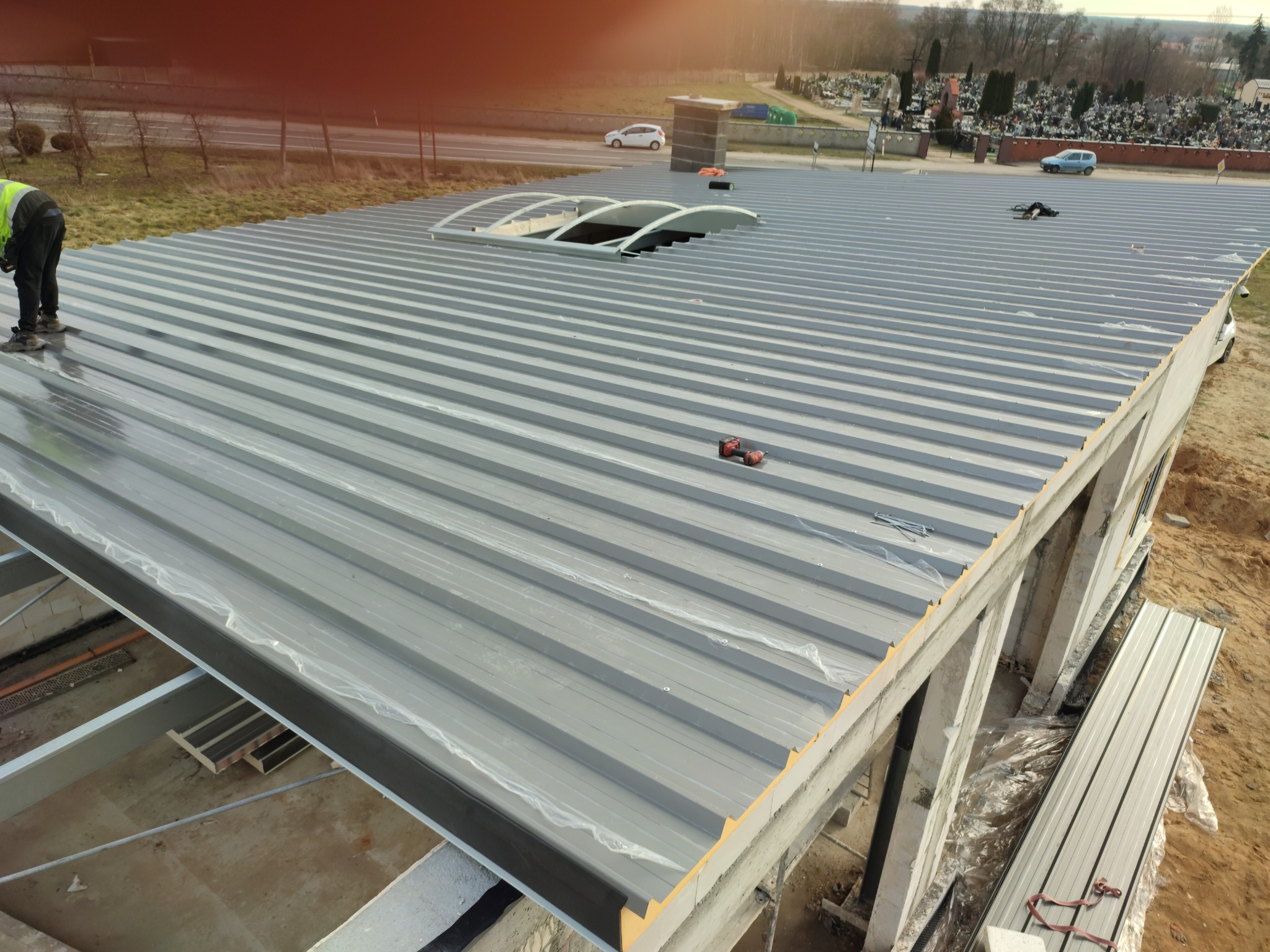 Budowa hali: Widok z góry na stalową konstrukcję dachu z świetlikiem łukowym w trakcie montażu, z pracownikiem w odblaskowej kamizelce i narzędziami na powierzchni, w tle cmentarz.