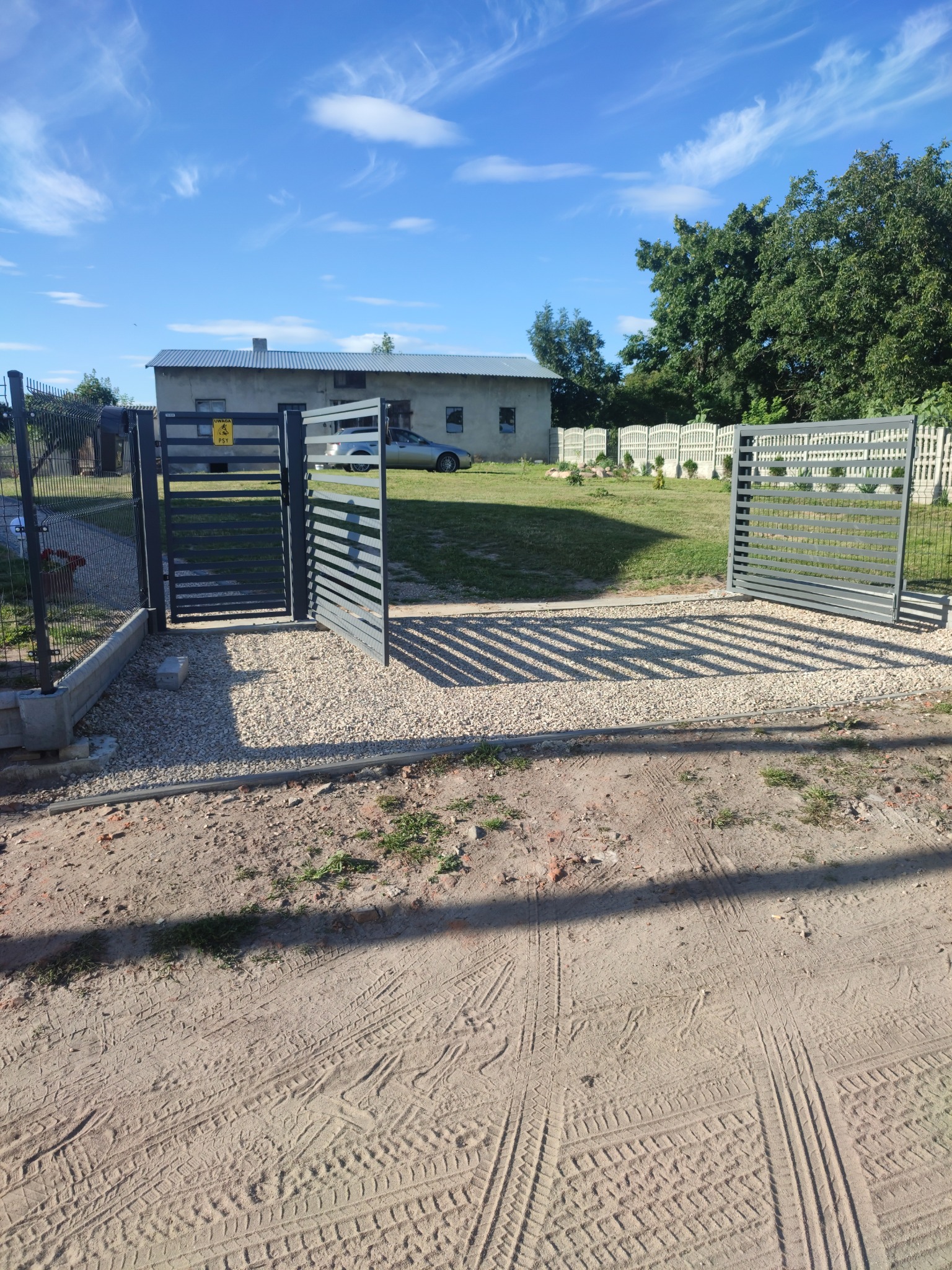 Nowoczesna, szara brama wjazdowa z poziomymi szczeblami, otwarta na posesję z domem i ogrodem, widoczna tabliczka ostrzegawcza.