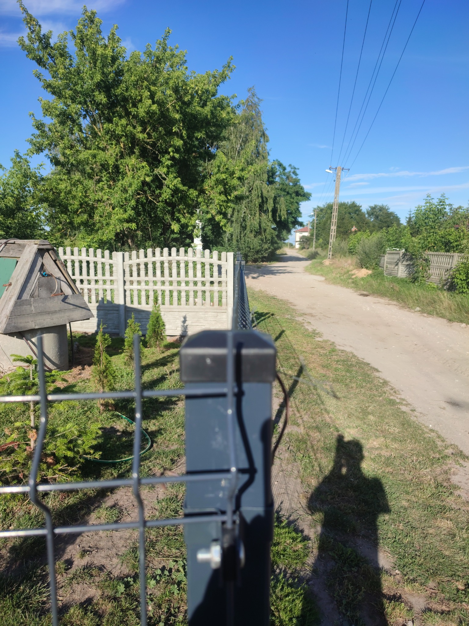 Fragment ogrodzenia panelowego z widocznym słupkiem, w tle betonowe przęsła, studnia z daszkiem, droga polna i cień osoby robiącej zdjęcie.