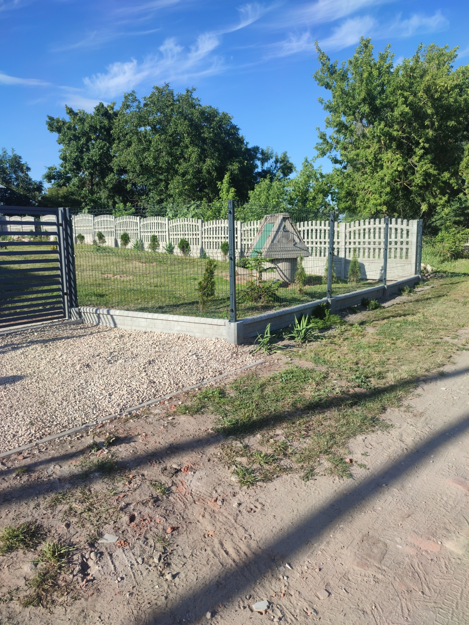 Nowoczesne ogrodzenie panelowe z bramą wjazdową, betonowa podmurówka, jasne żwirowe podłoże i studnia z drewnianą obudową w tle, widok w słoneczny dzień.