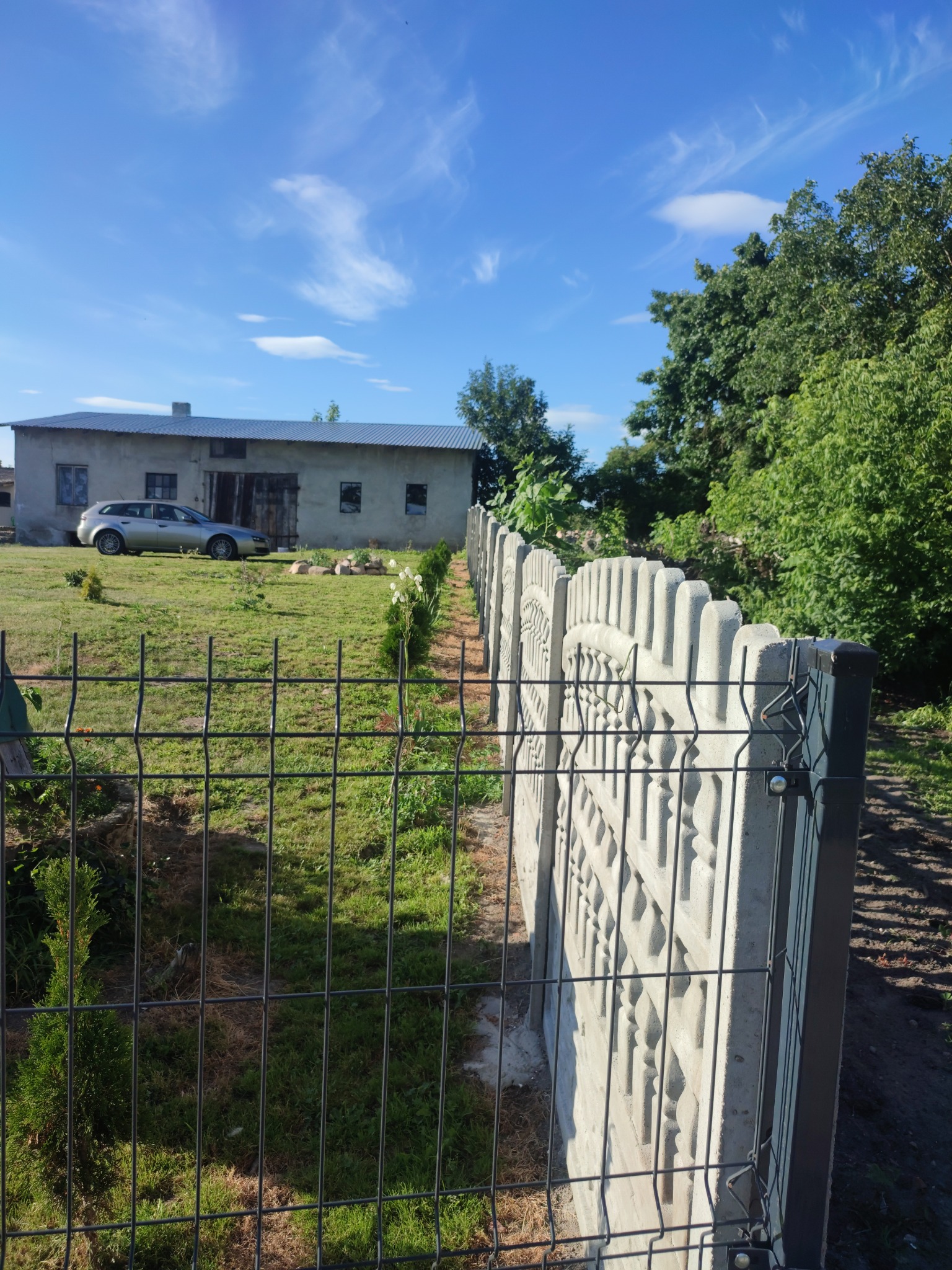 Fragment posesji z częściowo zamontowanym ogrodzeniem panelowym i betonowymi przęsłami dekoracyjnymi, widok na budynek gospodarczy i zaparkowany samochód w słoneczny dzień.