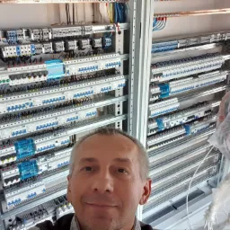 Selfie elektryka na tle rozdzielnicy z licznymi zabezpieczeniami, przewodami i listwami zaciskowymi, widoczne kable w osłonie.