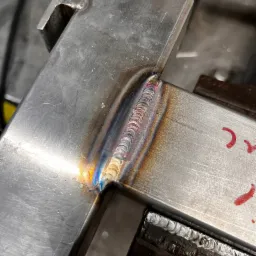 Spoina na stalowym profilu zamkniętym, widoczne tęczowe przebarwienia termiczne, całość mocowana w imadle ślusarskim.