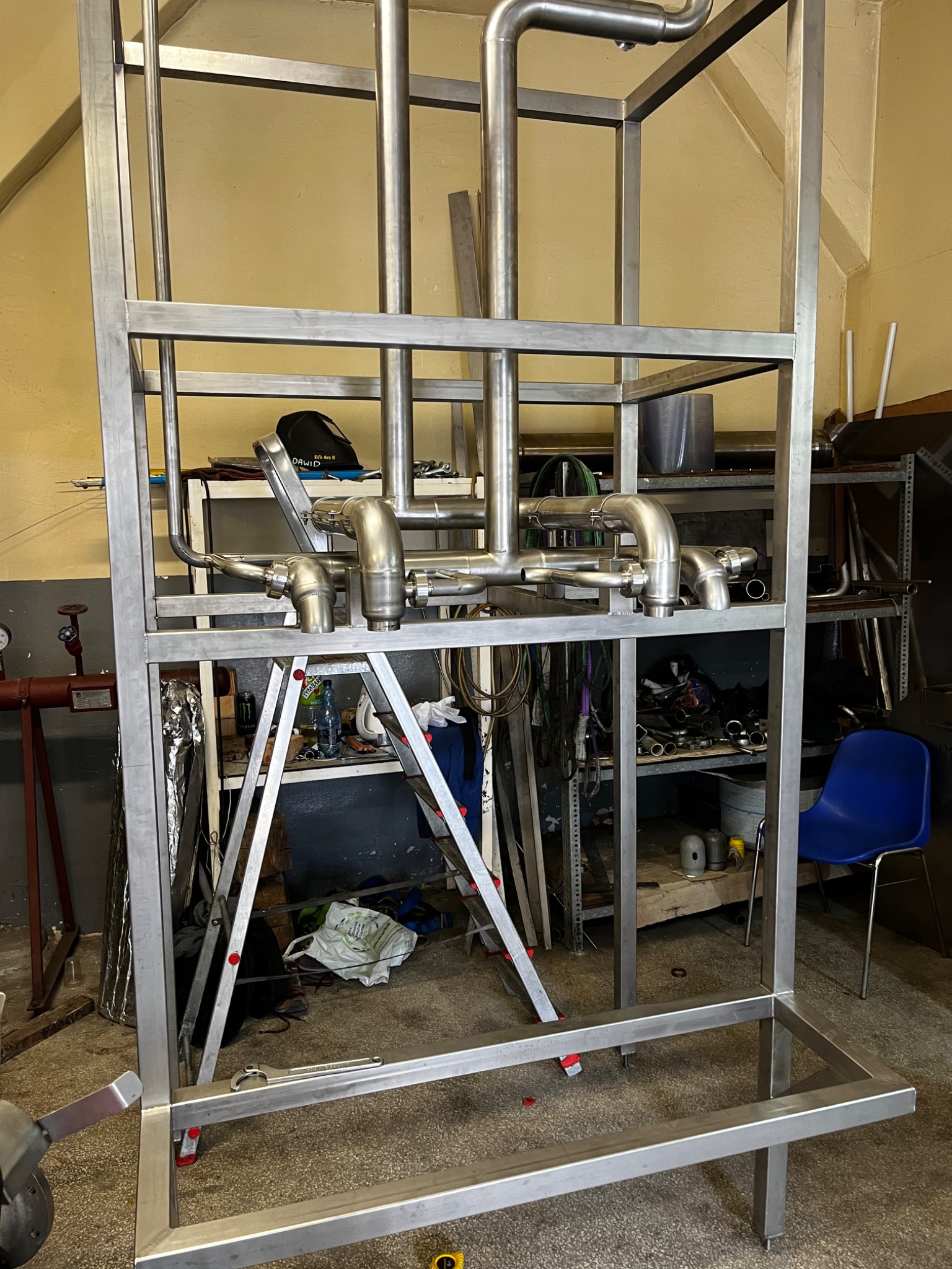 Stalowa konstrukcja z rurami i ramą na kółkach, prawdopodobnie element większej instalacji przemysłowej, widoczna drabina i narzędzia w tle.