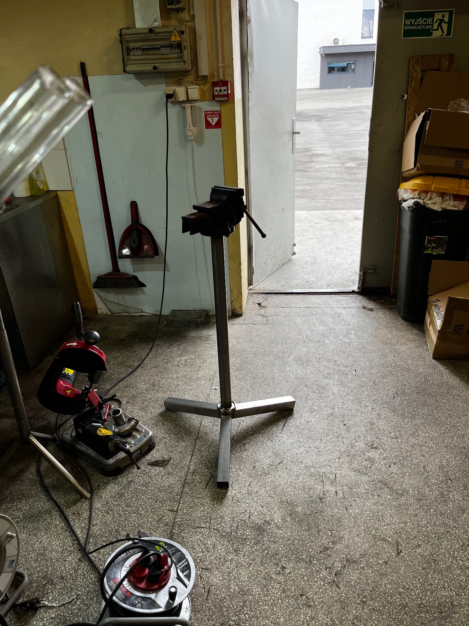 Warsztatowe ujęcie z bliska: stojak pod imadło z zamocowanym imadłem, piła taśmowa do metalu, przedłużacz zwinięty na bębnie, w tle otwarte drzwi do hali.