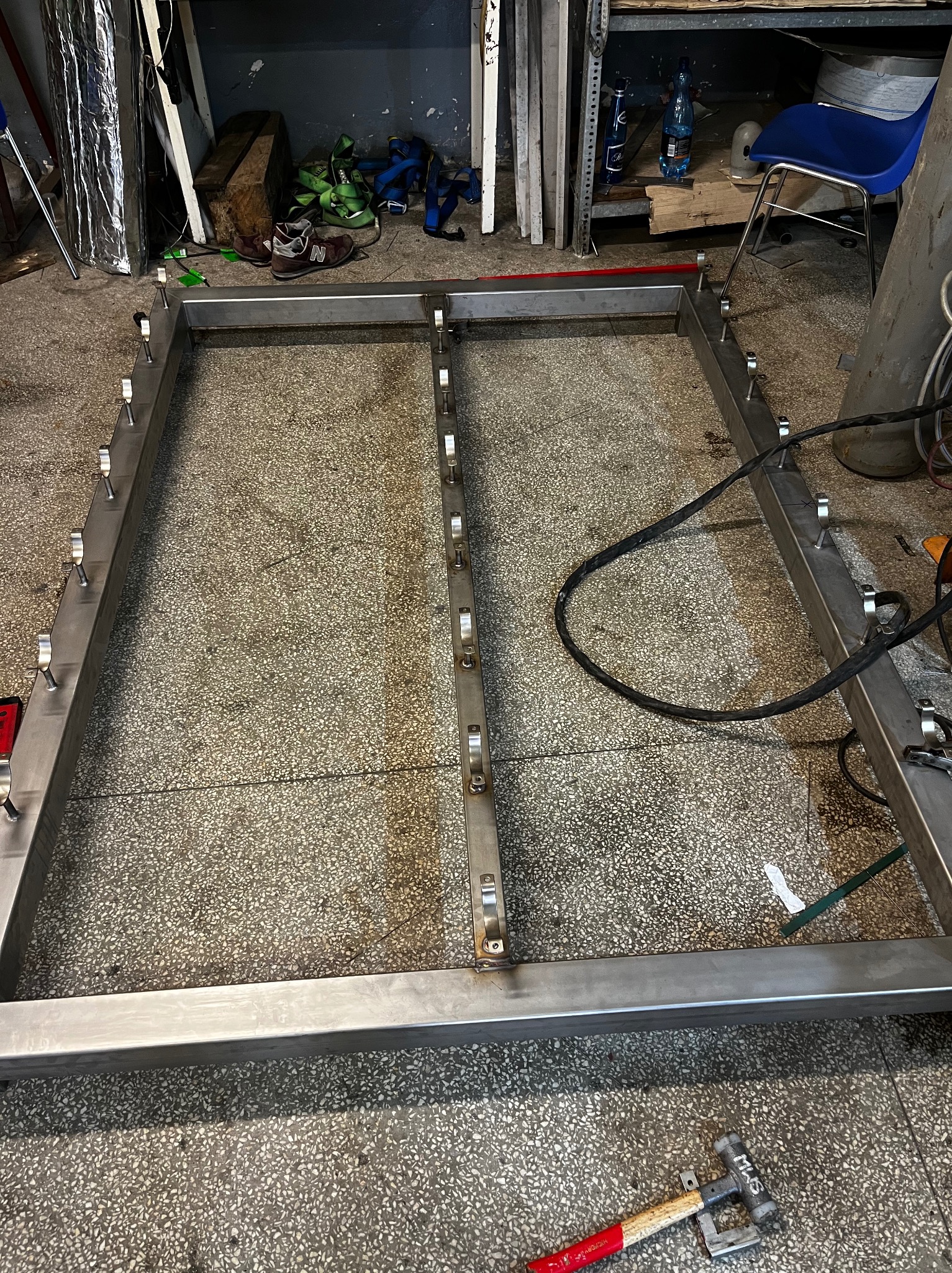 Stalowa rama konstrukcyjna z przyspawanymi uchwytami, widoczna w warsztacie na posadzce lastryko, z narzędziami i akcesoriami w tle.