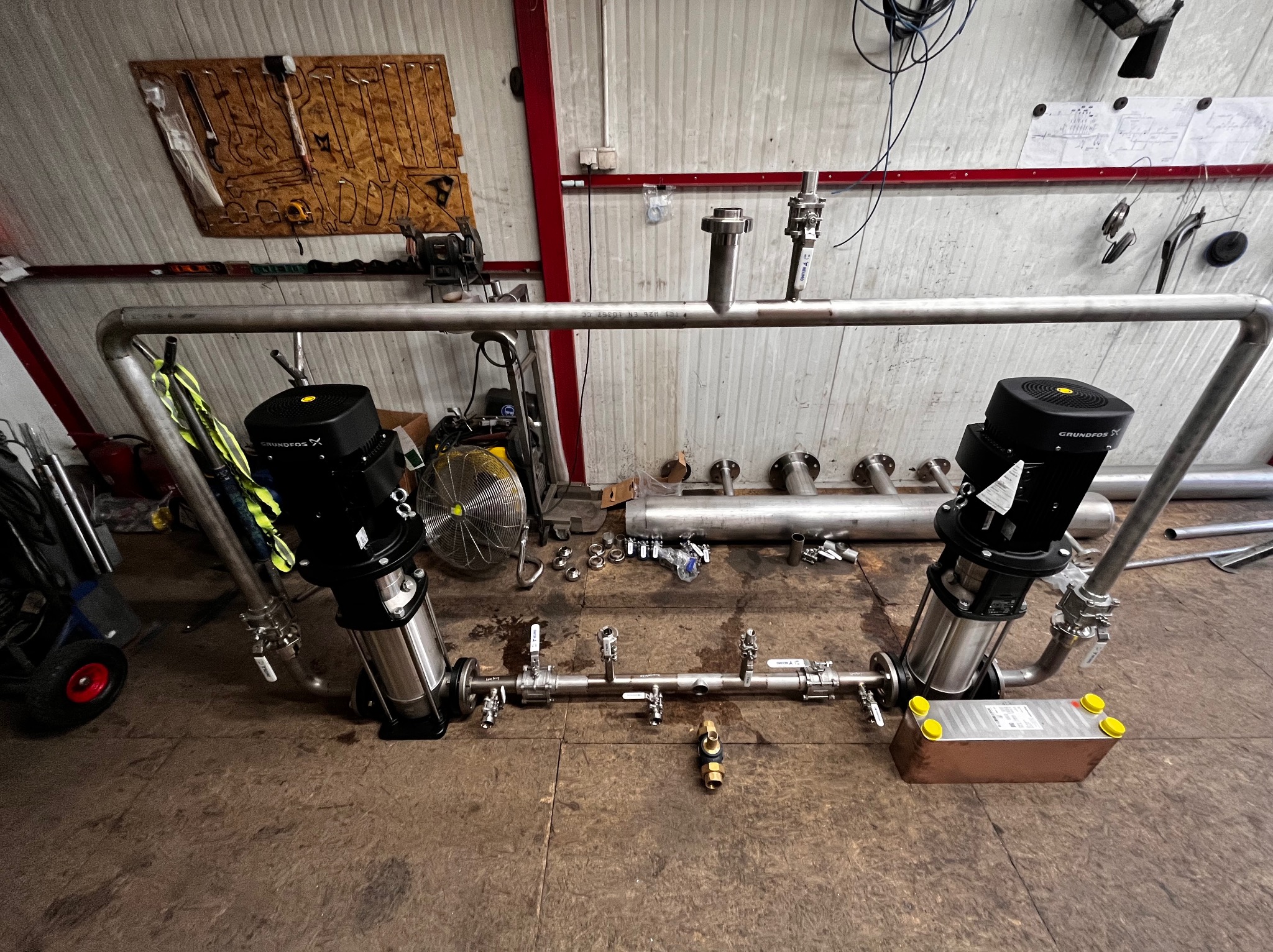 Konstrukcja z rur i pomp marki Grundfos, widoczne liczne złączki i zawory, na podłodze widoczne ślady po spawaniu, na ścianie wisi tablica z narzędziami.
